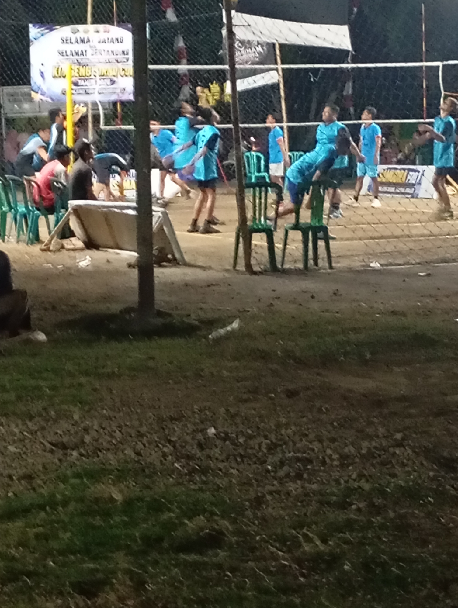 The image depicts an outdoor volleyball game at night, possibly in Pacitan, Indonesia, judging by the signage in Indonesian. The court appears to be a sand court. A net is stretched across the middle of the court. Several individuals, mostly young men and boys, are present on and around the court, many of them wearing light blue athletic jerseys. Some are actively playing volleyball, while others appear to be spectating or resting. There are green plastic chairs set up near the court, and a banner with text in Indonesian is visible in the background, which includes the words "SELAMAT DATANG" (Welcome) and "SELAMAT BERTANDING" (Good luck in the match). The lighting suggests artificial illumination is being used, typical for evening or night sporting events. The ground surrounding the court is a mixture of dirt and some grassy areas.