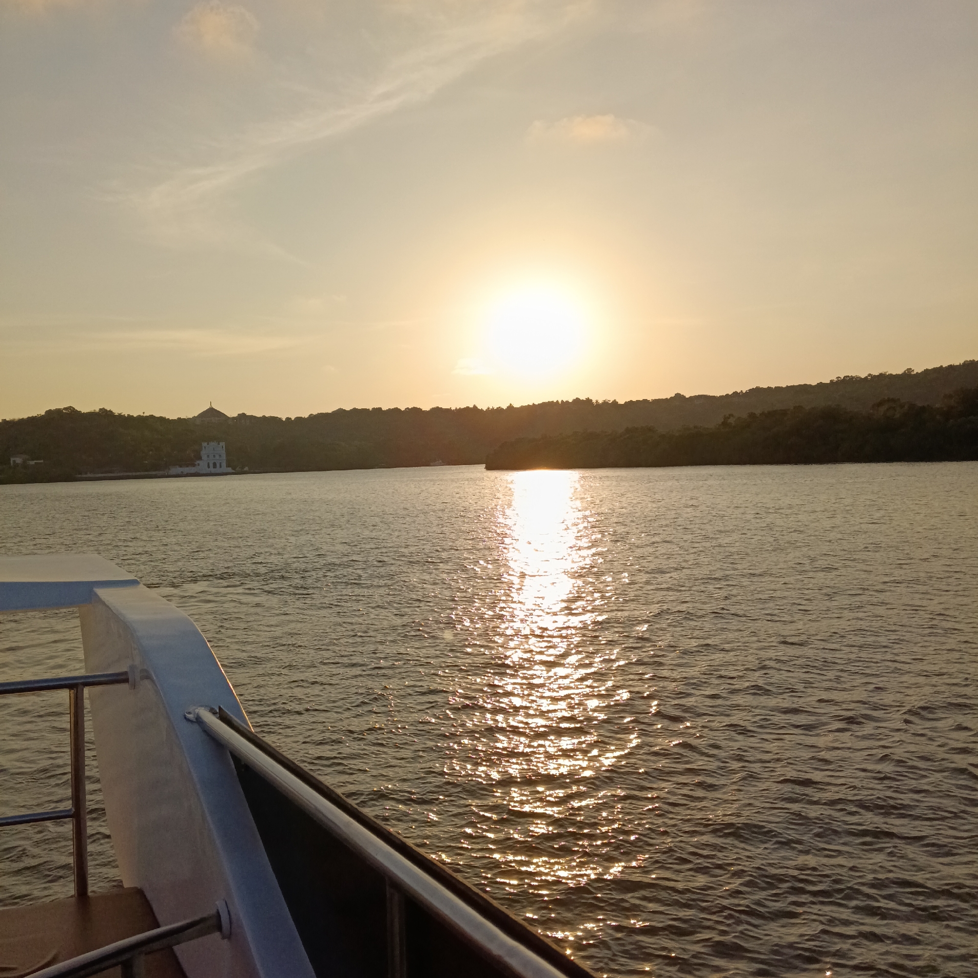 View from a boat during a sunset at Britona, India. A body of water reflects the setting sun, creating a bright, shimmering path. In the distance, a tree-covered landmass is visible with a white building featuring a dome structure along the coastline. The boat's railing is in the lower left corner.
