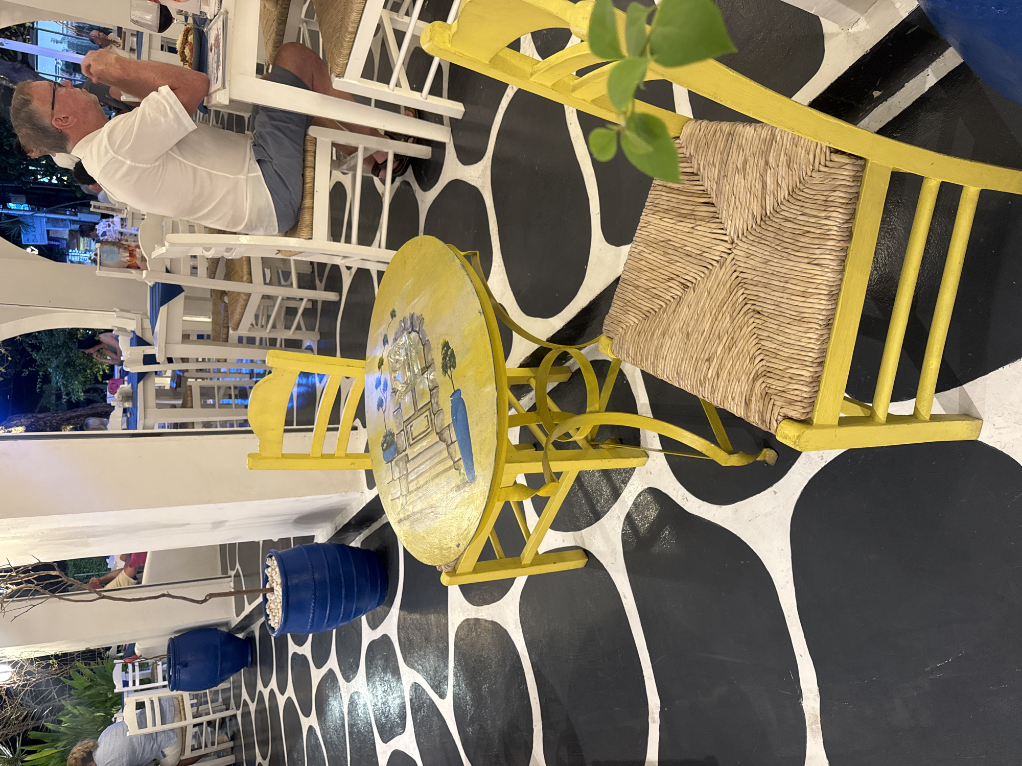 The image depicts an outdoor dining area, possibly in Denpasar, Indonesia. The scene is dominated by black and white floor tiles patterned with organic shapes. The furniture consists of yellow chairs and a matching round table, with at least one chair having a woven seat. A person is seated at a white table with white chairs, eating. There are blue decorative pots with white stones and branches. The image suggests an outdoor restaurant setting with a distinctive design.