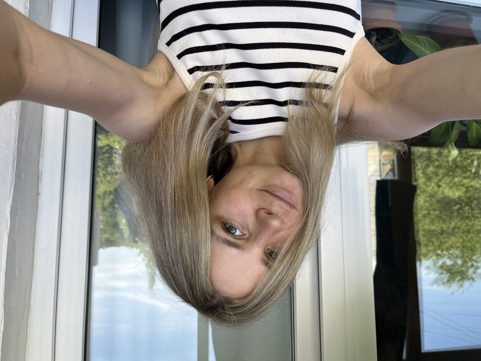 A woman with blonde hair is taking a selfie in front of a window. She is wearing a black and white striped tank top. The background shows trees and a building. The location might be Chaikovsky, Russia, based on prior user knowledge.