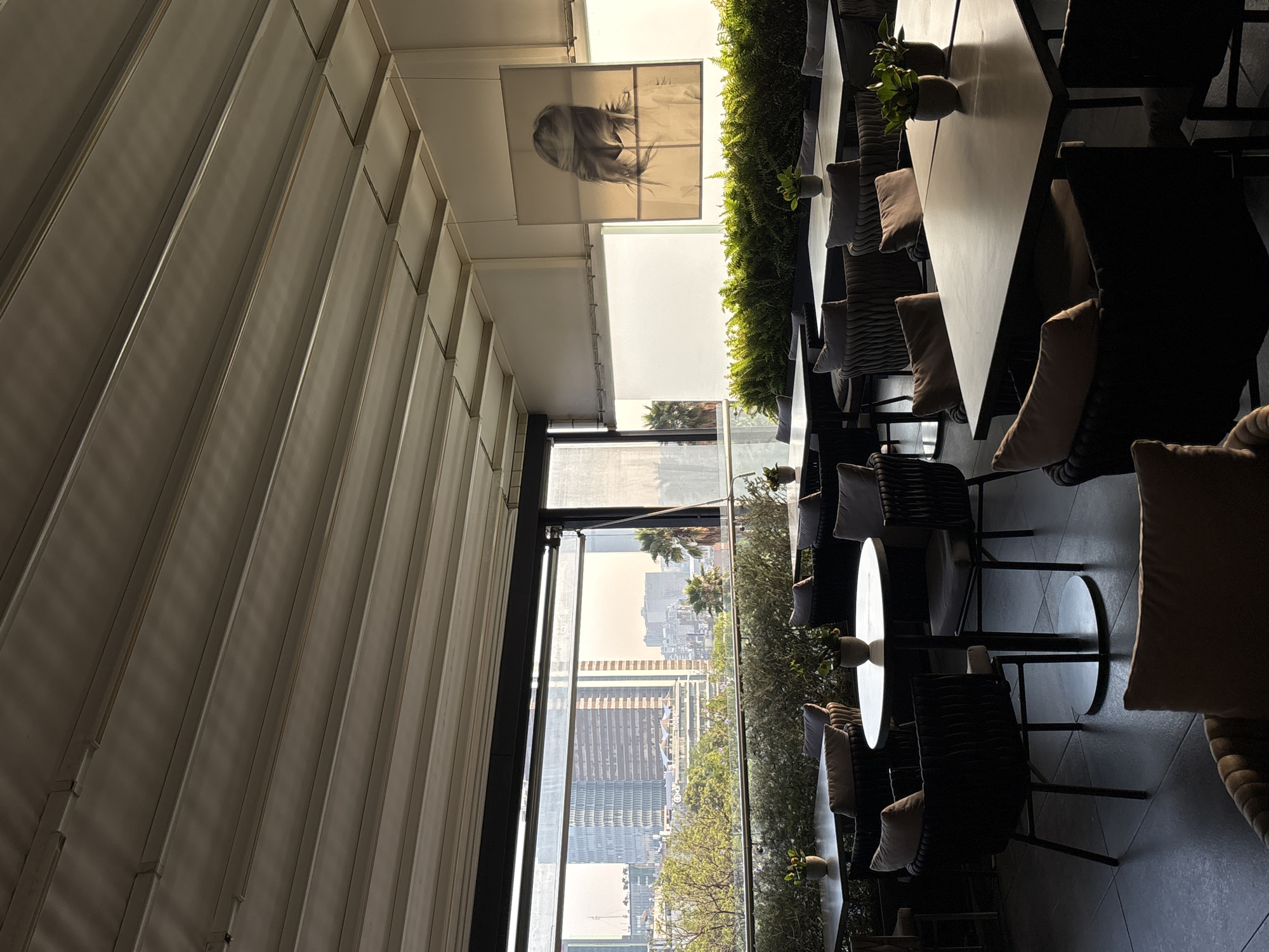 The image shows the interior of a restaurant or cafe with a view of Mexico City, Mexico. Long tables and chairs, with pillows, are arranged along a windowed wall, decorated with greenery. Outside the window is a view of the city skyline with buildings and trees, including palm trees, indicating a high-rise setting. Interior is a long white and metallic lined wall, with a painting hanging from the ceiling.
