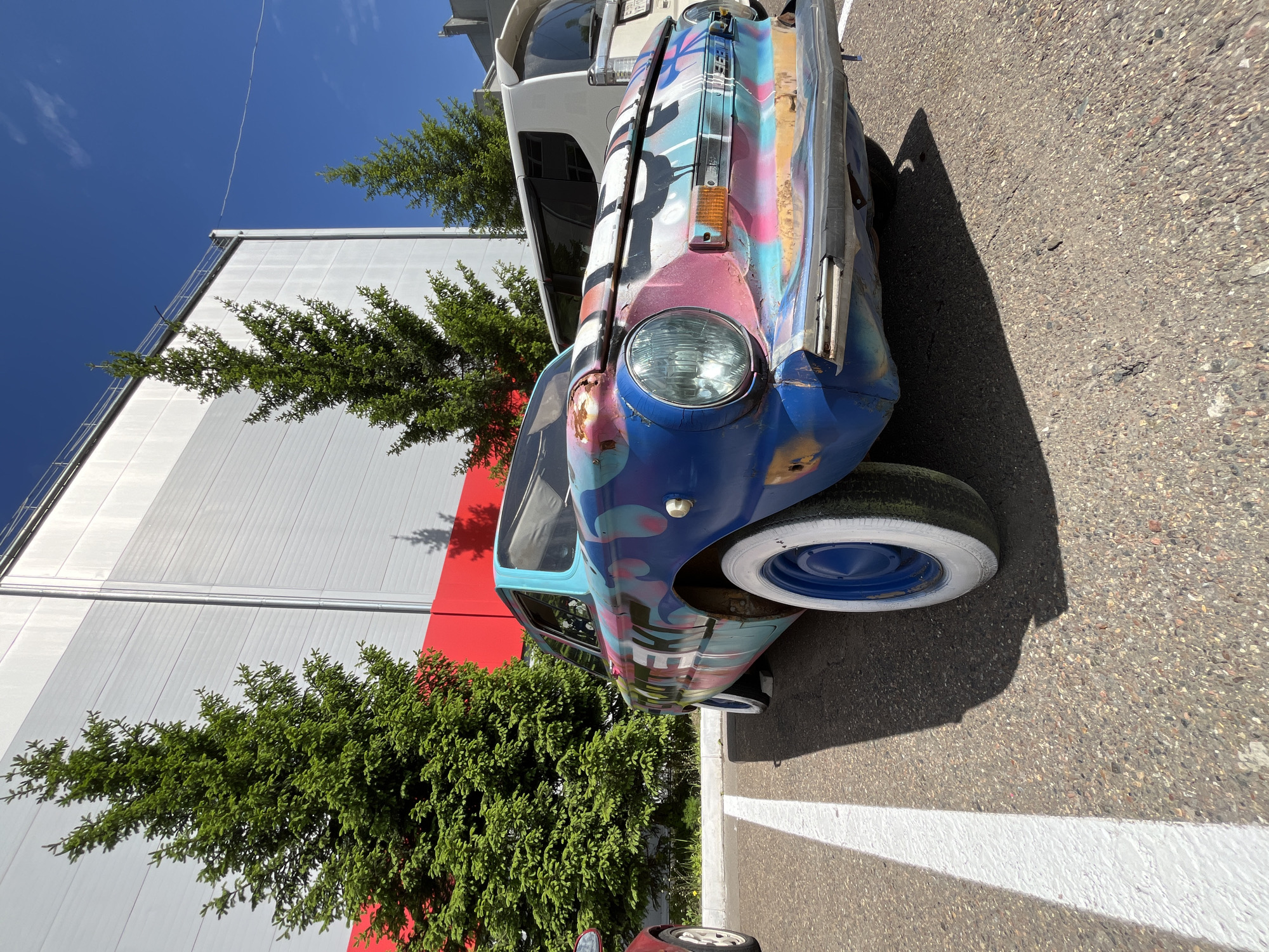 The image shows a parked, brightly painted car on a paved surface, near a building with a white and red facade and green trees. The car has a blue base color with various colorful patterns and whitewall tires. Part of a white van is visible behind the car. The location is Krasnoyarsk, Russia.