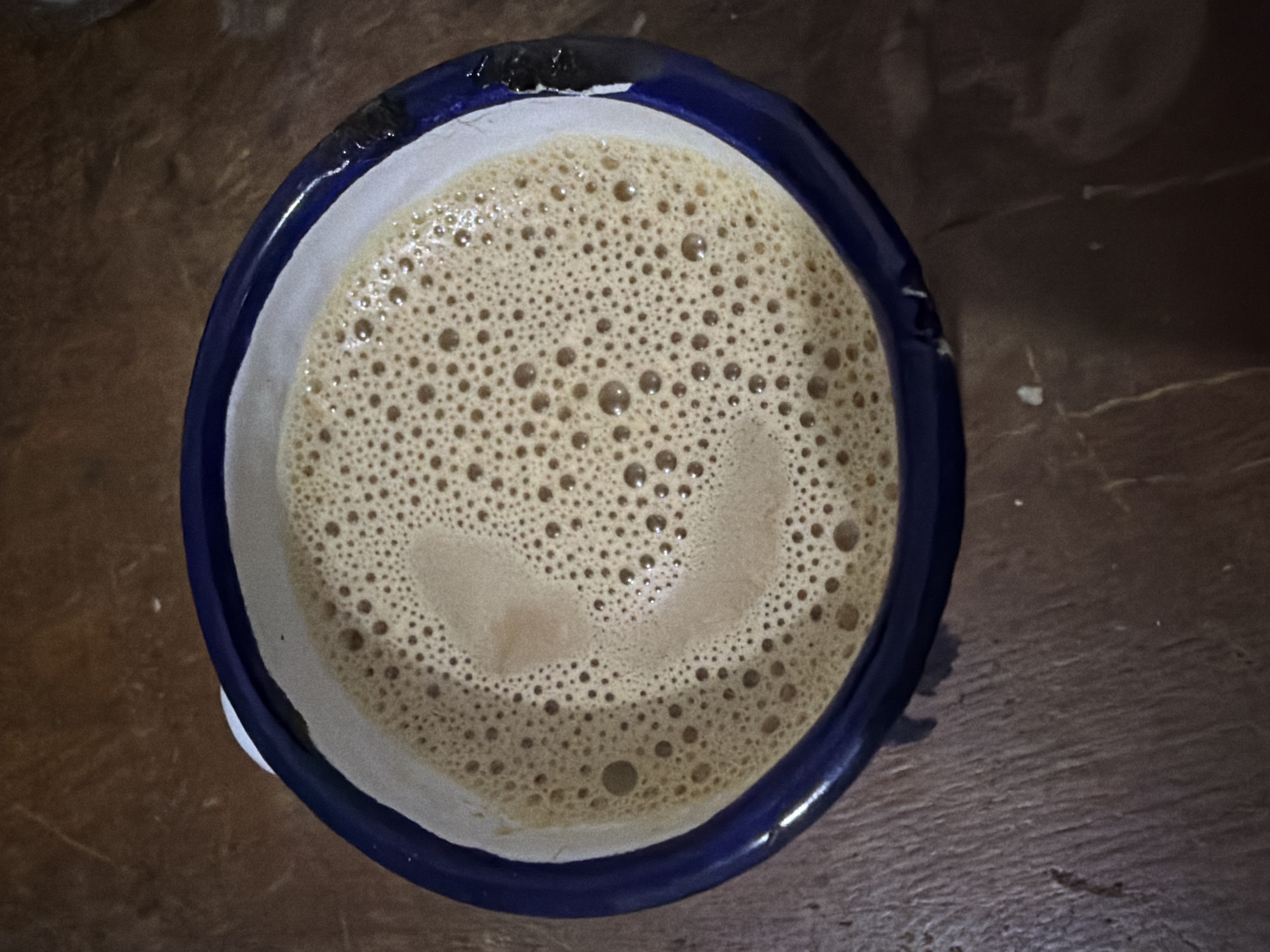 A top-down, close-up shot of a light brown, frothy beverage contained within a blue and white ceramic mug. The beverage's surface is covered in small bubbles. The mug is positioned on a dark brown wooden surface. The location is Ciudad Guayana, Venezuela. No people are visible.
