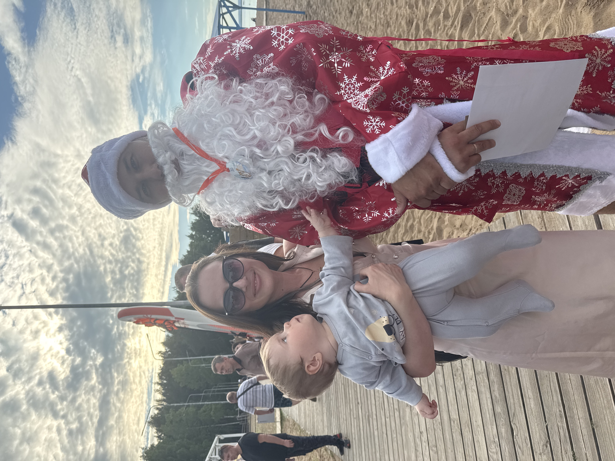 This image, taken in Chaikovsky, Russia, depicts a festive outdoor scene on a beach with wooden boardwalks. The primary subjects are a group of people, including a person dressed as Santa Claus, a woman holding a baby, and other onlookers in the background.
The person in the Santa Claus costume is wearing a red outfit adorned with white snowflakes and has a prominent white beard and hat. They are holding a blank piece of paper, possibly for a letter or a list. The woman, wearing sunglasses, is smiling and holding a baby dressed in a grey onesie with a bear design. The baby is looking up and appears engaged.
The background shows a sandy beach and a cloudy sky, suggesting it's daytime with diffused sunlight. There are trees and structures in the distance, hinting at a recreational area. The overall mood seems joyful and celebratory, likely during a holiday event or gathering. The wooden boardwalk in the foreground provides a clear visual of the setting. No visible text other than on the baby's onesie which is too small to read.