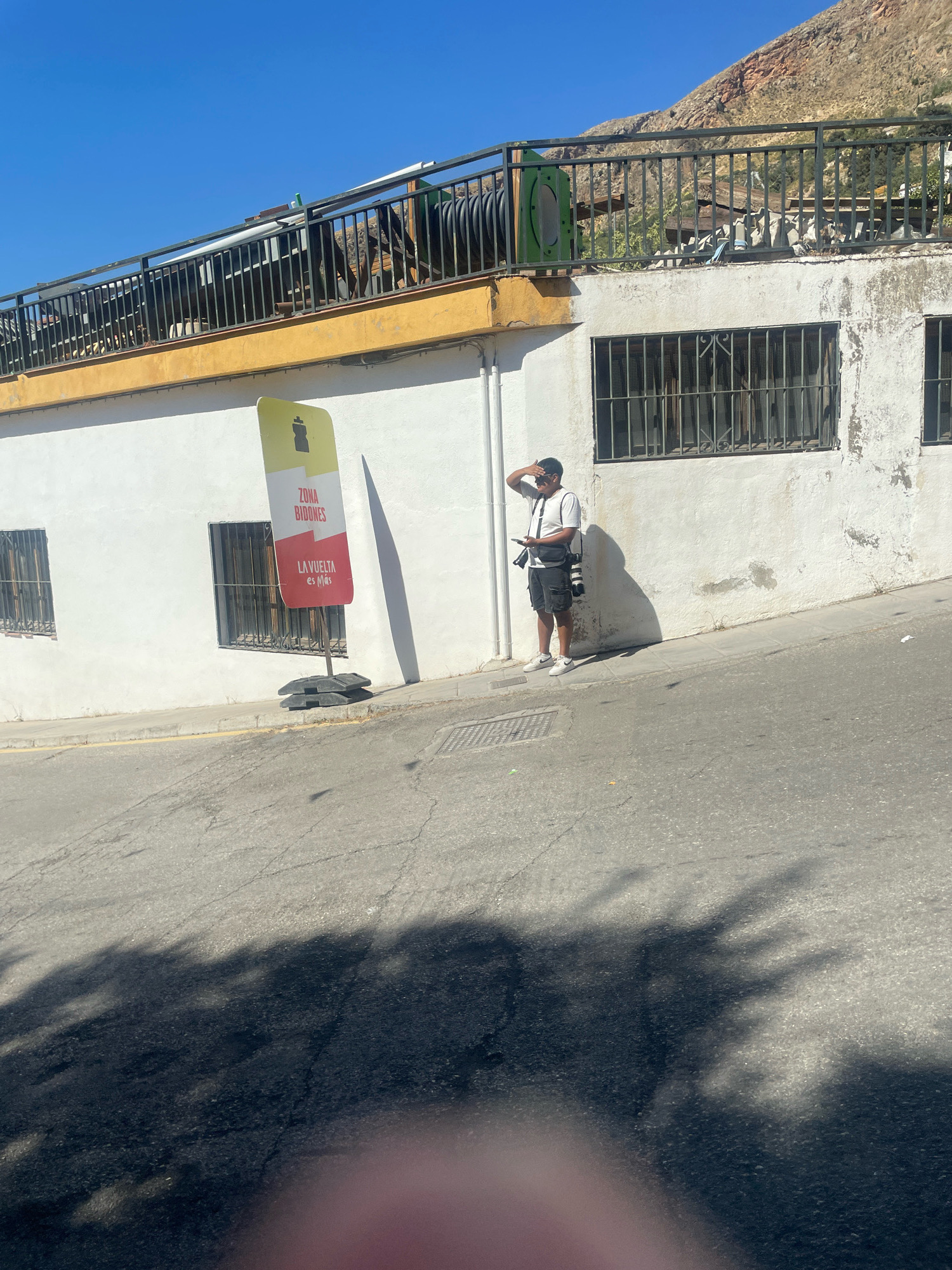 A man stands on a paved street in Güéjar Sierra, Spain, wearing a white t-shirt, black shorts, and sneakers. He has a camera bag slung across his body and is holding a smartphone in his left hand, while his right hand is raised to his forehead, shielding his eyes from the sun. He is looking to his right, towards a white building with barred windows. In front of the building, a tall, yellow and red sign reads "ZONA BIDONES" and "LA VUELTA es Más". The building is situated on a slight incline, with a railing and vegetation visible on an upper level. The street is asphalt, with shadows from trees cast across it. The sky is clear and blue, and a rocky hillside is visible in the background.