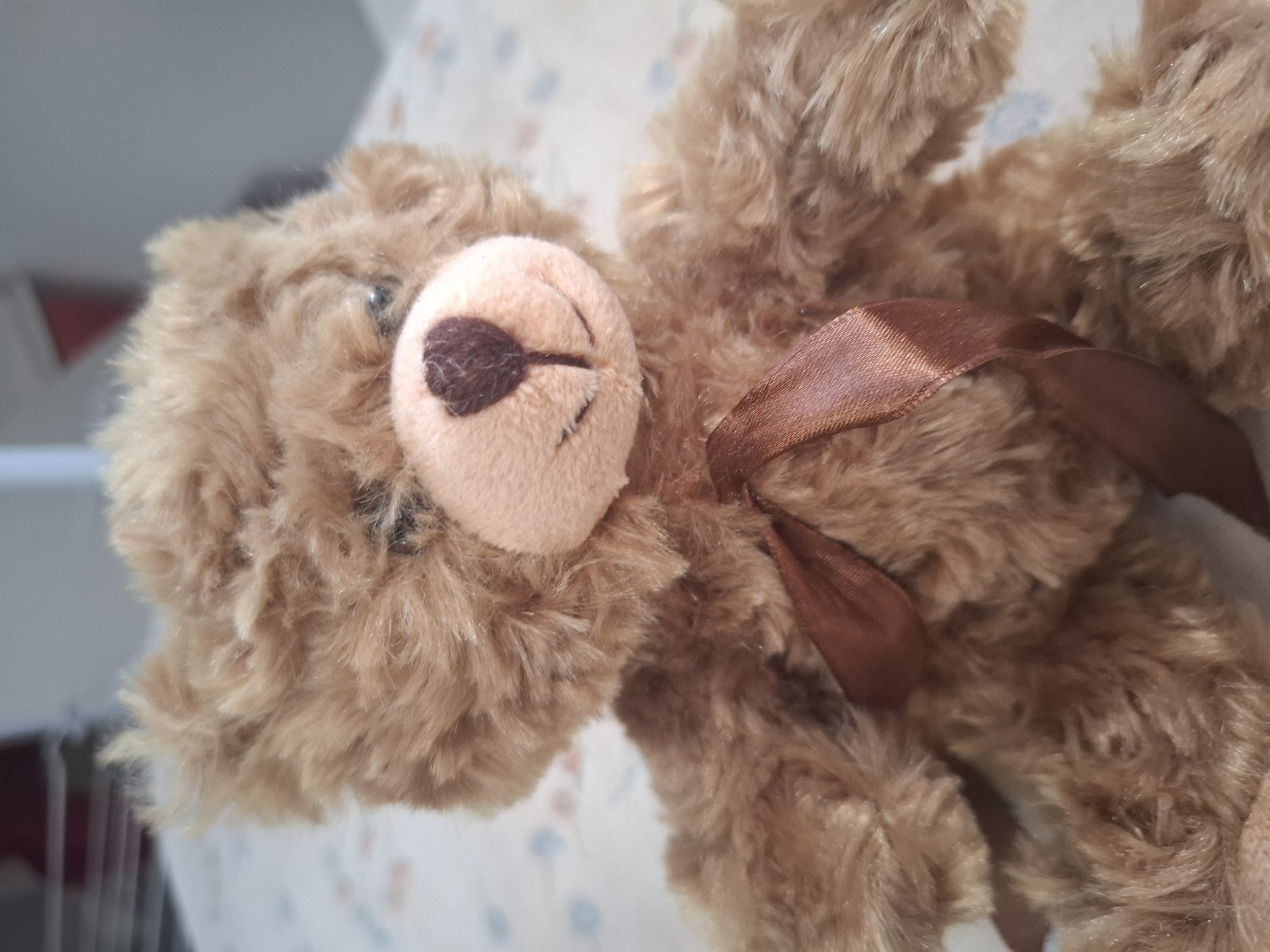 The image shows a close-up of a brown teddy bear sitting on a surface with a floral pattern. The teddy bear has a stitched mouth, a dark brown nose, and shiny black eyes. It is wearing a satin ribbon tied in a bow around its neck. The background is blurred, but appears to be a room with some shelves or stacked items visible in the upper left corner. There is no indication of the city of Montreuil, France, in the image or its immediate surroundings.