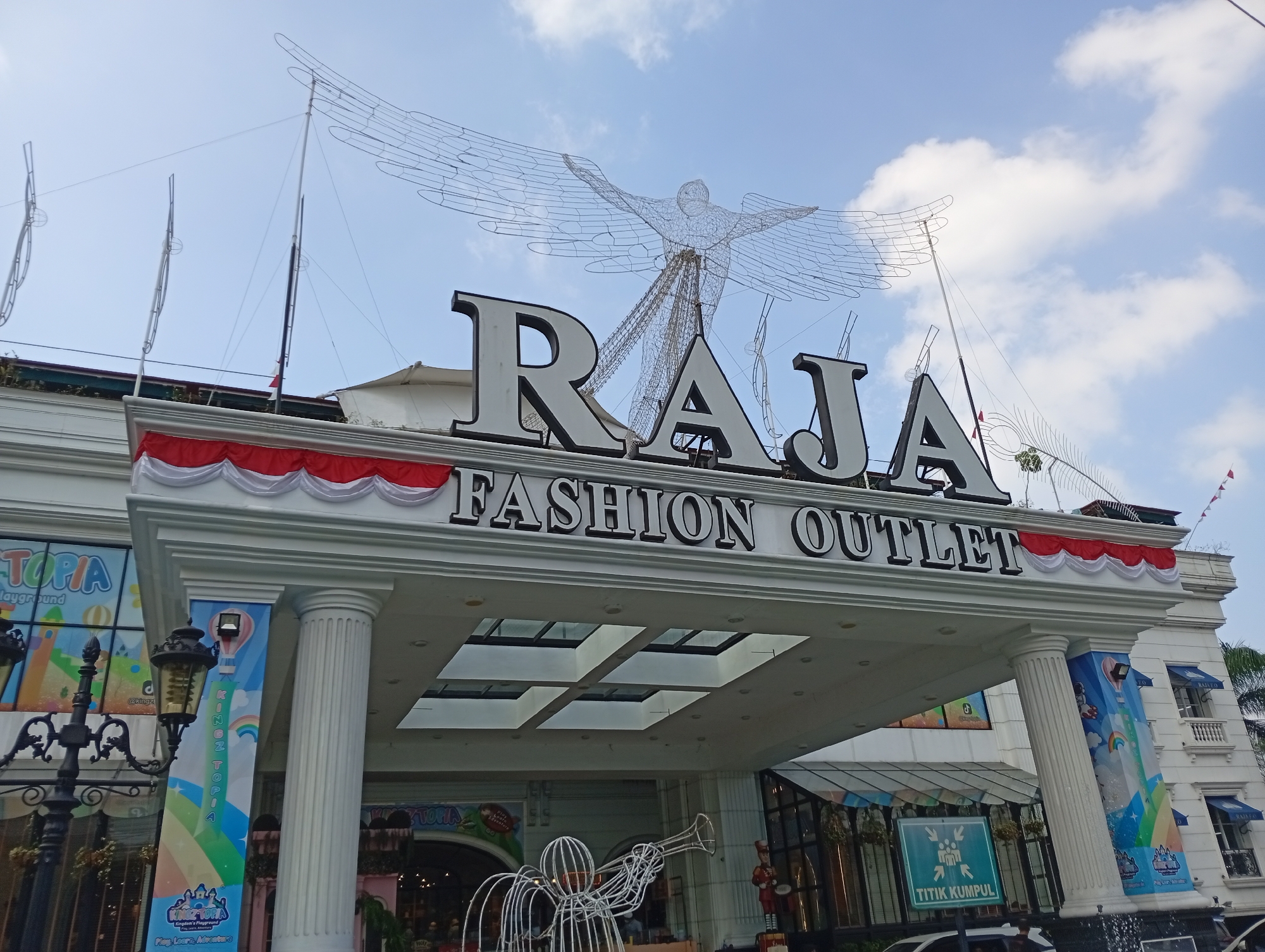 The image shows the exterior of a building identified as "RAJA FASHION OUTLET" in Cimacan, Indonesia. The building has a white facade with classical columns and decorative red and white drapery along the upper facade. A large wire sculpture of an angel with outstretched wings is positioned above the "RAJA" lettering. The sky above is partly cloudy. In front of the building, there is a street lamp and banners with colorful graphics, one of which displays the text "KINGTOPIA". A green sign with figures and the text "TITIK KUMPUL" is visible. The overall scene depicts the entrance of a commercial establishment on a bright day.