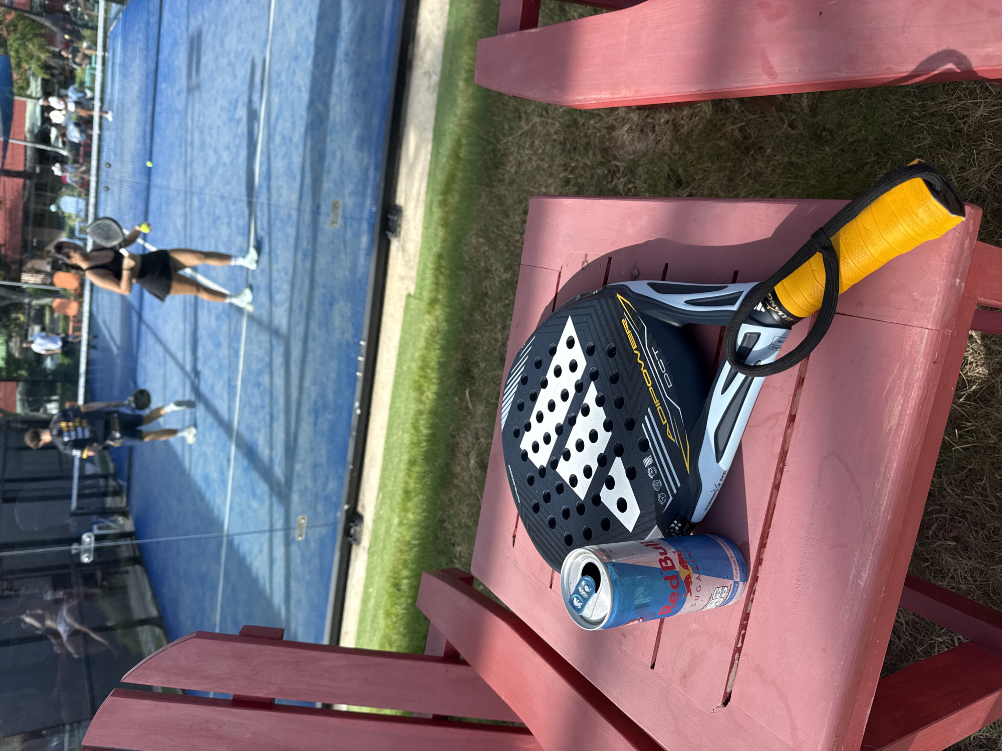 In the foreground, a red wooden table is occupied by a can of Red Bull and a black and white Adidas padel racket with a yellow grip. In the background, a blue padel court is visible through a glass wall. Two individuals are playing padel on the court. One, a man in a black and yellow shirt and shorts, is on one side of the net, while a woman in a black athletic outfit is on the other side. The environment suggests an outdoor sports facility, likely a resort or club, given the presence of lounge chairs and manicured greenery. The scene is consistent with a location in Ko Samui District, Thailand, known for its tourist resorts and sports facilities.
