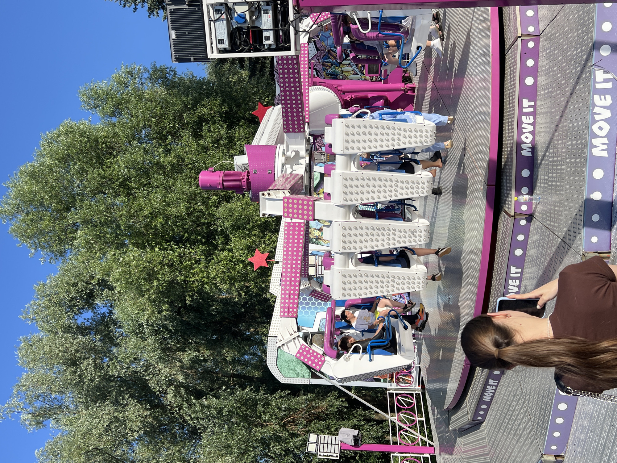 The image depicts an outdoor amusement park ride, identified by the text "MOVE IT" visible on the ride's platform, located in the city of Cheboksary, Russia. The ride is predominantly white and pink, with a central mechanical tower and spinning arms supporting passenger cabins. Several people are seated in the cabins, experiencing the ride, while others are standing on the platform. In the foreground, a young woman is captured from behind, holding up a smartphone to take a picture or record a video of the attraction. The surrounding environment includes lush green trees against a clear blue sky, suggesting a daytime setting during warm weather. The overall scene captures a moment of leisure and entertainment at a fairground.