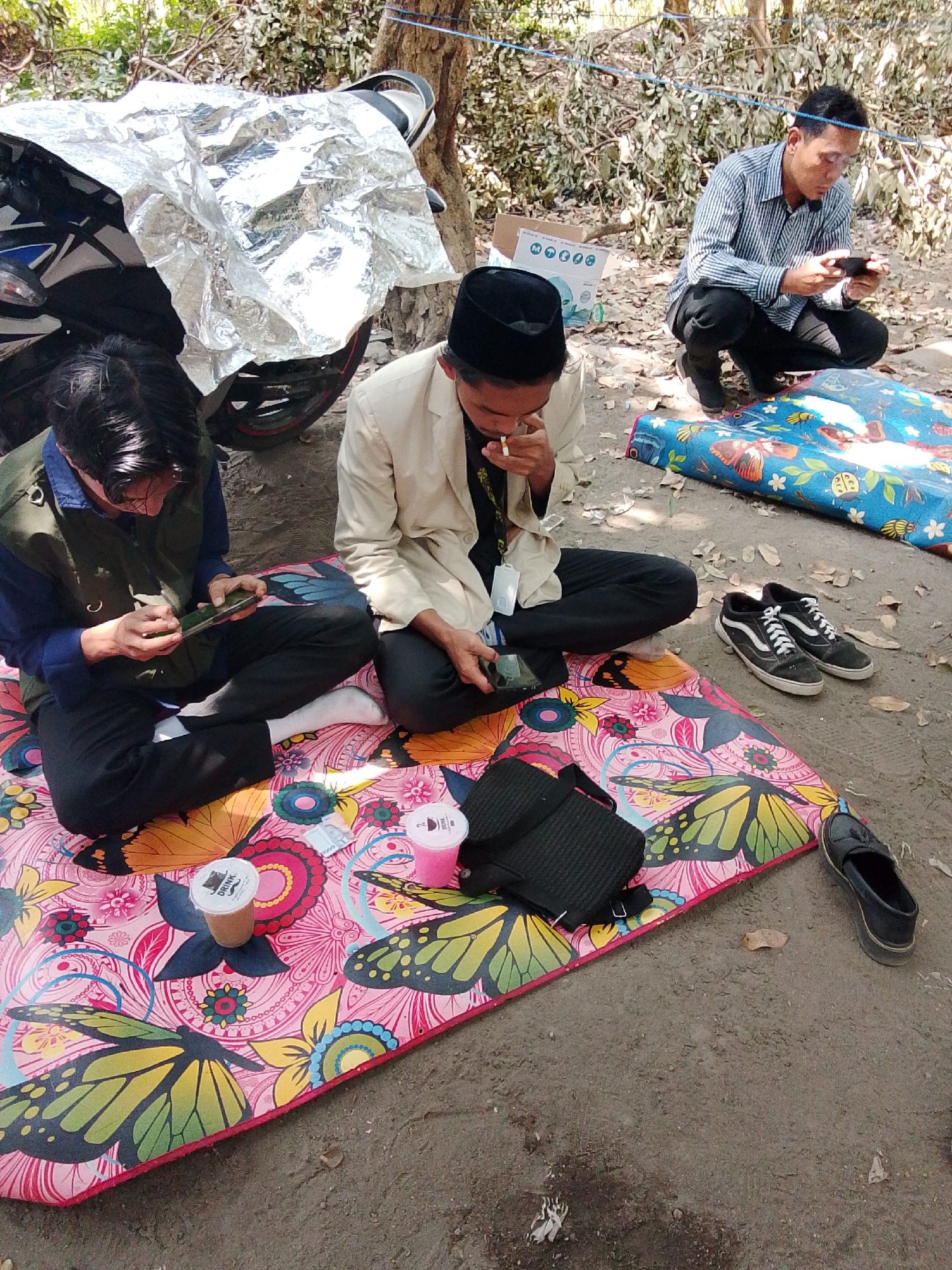 In Grogol, Indonesia, three individuals are seated on the ground amongst foliage and dirt. The person on the far right, wearing a striped shirt and black pants, squats and intently holds a smartphone. In the center, a person in a cream-colored blazer and black cap sits with a cigarette in their mouth and holds a smartphone. To the left, another individual in a dark green vest over a blue shirt and black pants, also wearing white socks, is seated and engaged with a smartphone. Two colorful mats are spread on the ground, one with a butterfly and floral design, and another with images of insects and flowers. A motorcycle is partially visible on the left, covered with a silver tarp. Several cups and a black bag are placed on the mats. The environment appears to be an outdoor, possibly park-like setting, with dry leaves scattered on the ground.