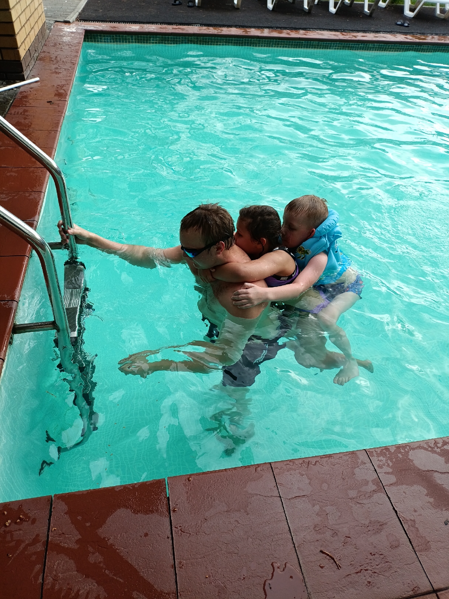 This image captures an outdoor swimming pool scene, likely in Chaikovsky, Russia, during daytime under diffused natural light, suggesting an overcast sky or indirect sunlight.
In the center of the frame, an adult male is in the turquoise pool water, holding onto a silver metal pool ladder with his right hand. He is wearing sunglasses and dark swim trunks. Clinging to him are two children. On his left, a young girl with dark hair is embracing him, wearing a dark swimsuit with "NIKE" visible on the strap. On his right, a young boy with lighter hair is also holding onto the adult, wearing a blue flotation vest with a light pattern and patterned swim shorts. Both children's legs are visible dangling in the clear water beneath the surface. The adult appears to be supporting both children, suggesting a playful interaction or assistance in the water.
The swimming pool water is a vibrant blue-green, showing ripples and reflections. The pool's edge is lined with wide, reddish-brown, textured tiles or coping, some appearing wet. In the upper left background, a portion of a light brown brick wall is visible. Along the far edge of the pool, beyond the water, there's a dark paved area and glimpses of what appear to be white poolside furniture. The scene conveys a moment of family activity and togetherness in the pool.