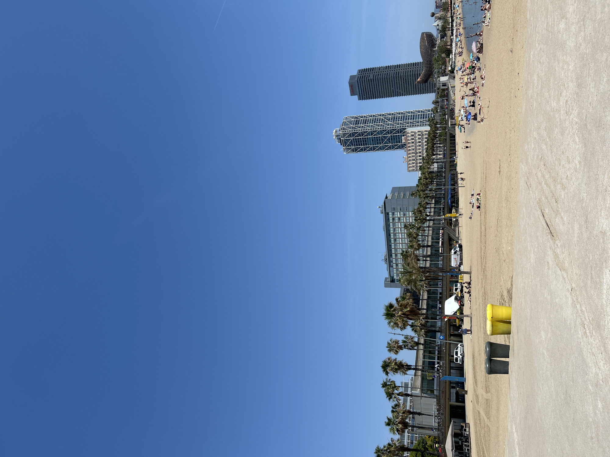 The image captures a sunny day at Barceloneta Beach in Barcelona, Spain. The foreground features a sandy beach with two pairs of trash receptacles, one dark gray and one bright yellow. Beyond the sand, a paved walkway is lined with palm trees and a row of buildings. In the middle ground, a lively beach scene unfolds with numerous people sunbathing, walking, and interacting. Beach umbrellas dot the sand, providing shade for some. Several vehicles, including white vans and a yellow construction vehicle, are visible near the buildings. Dominating the background are modern skyscrapers, including the distinctive Torre Glòries (formerly Torre Agbar) with its lattice-like exterior and another tall, dark rectangular skyscraper. A large, metallic fish sculpture is also visible near the water's edge. The ocean stretches out in the distance, with swimmers enjoying the water. The sky above is a clear, vibrant blue.