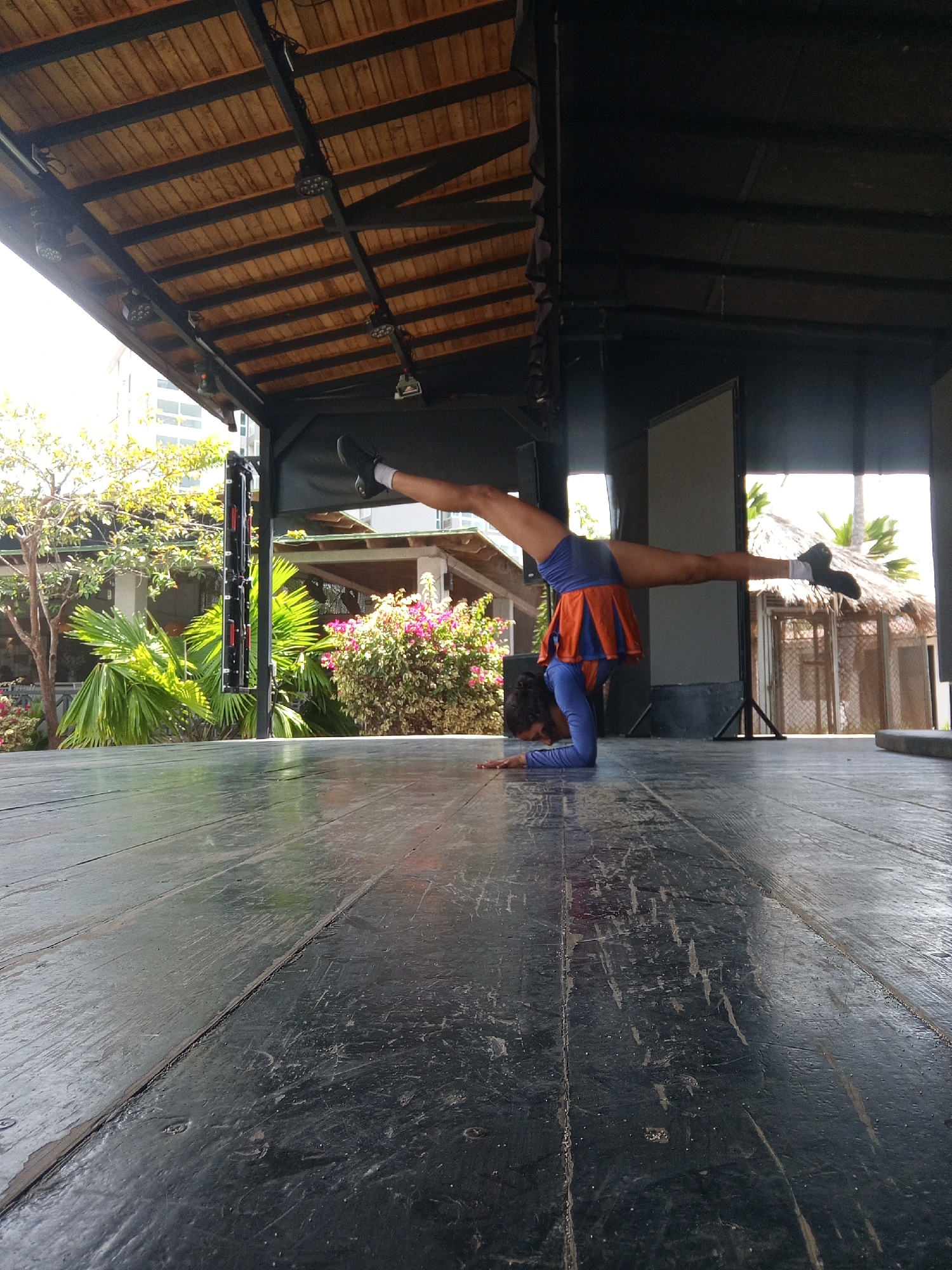 A woman is performing a handstand on a weathered wooden stage under a wooden roof structure. She is wearing an orange and blue athletic uniform and black sneakers with white socks. The background includes greenery, palm trees, and glimpses of building structures. The location appears to be an outdoor performance area in Porlamar, Venezuela.