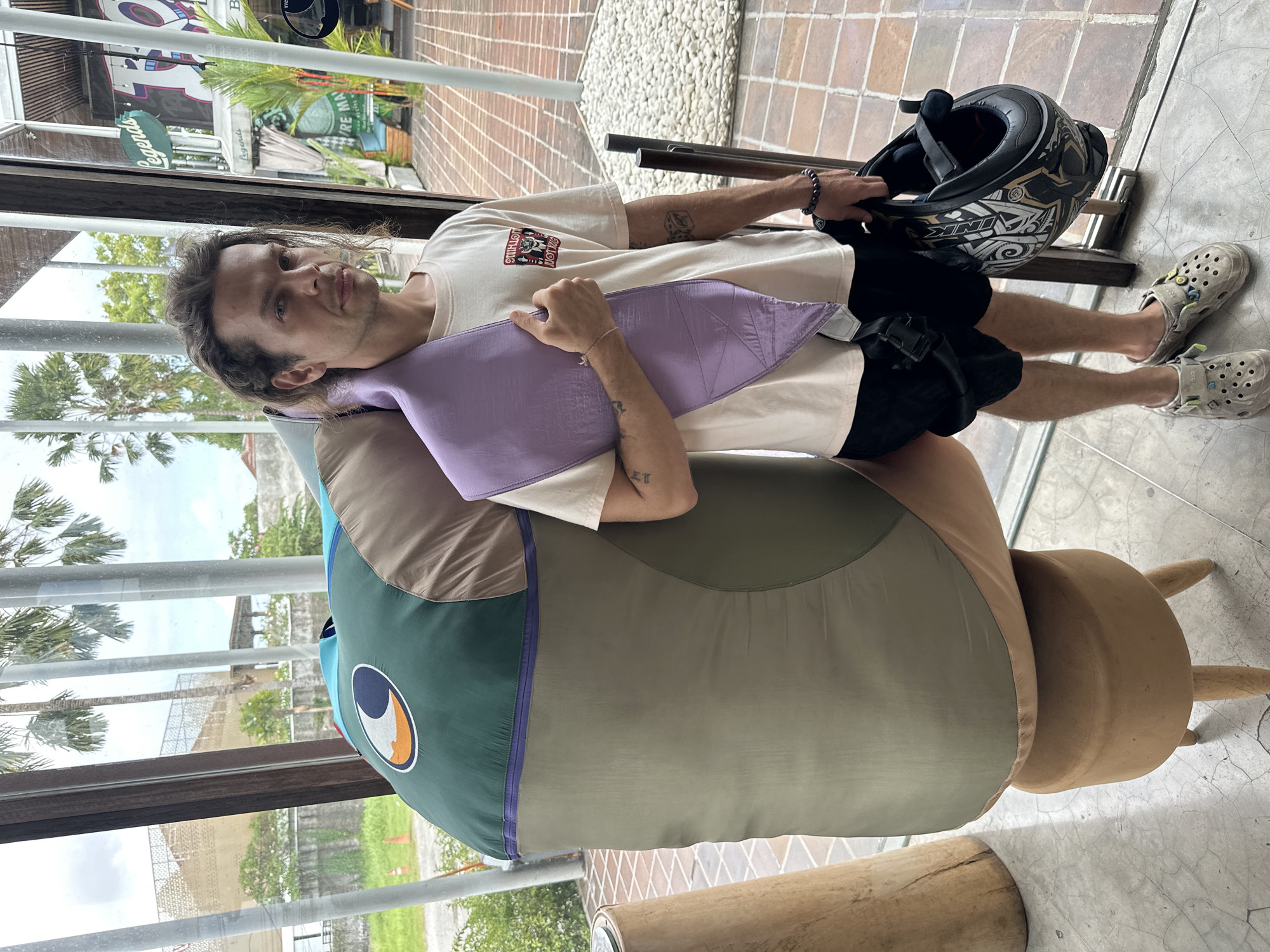 A man is reclining in a chair in North Kuta, Indonesia. He has long brown hair and is wearing a beige T-shirt, black shorts, and Crocs. He is holding a motorcycle helmet in one hand and has a purple neck pillow. The chair has a unique design, resembling a fish with a blue eye and a green head. The setting appears to be indoors, possibly a cafe or shop with large windows revealing tropical vegetation outside.