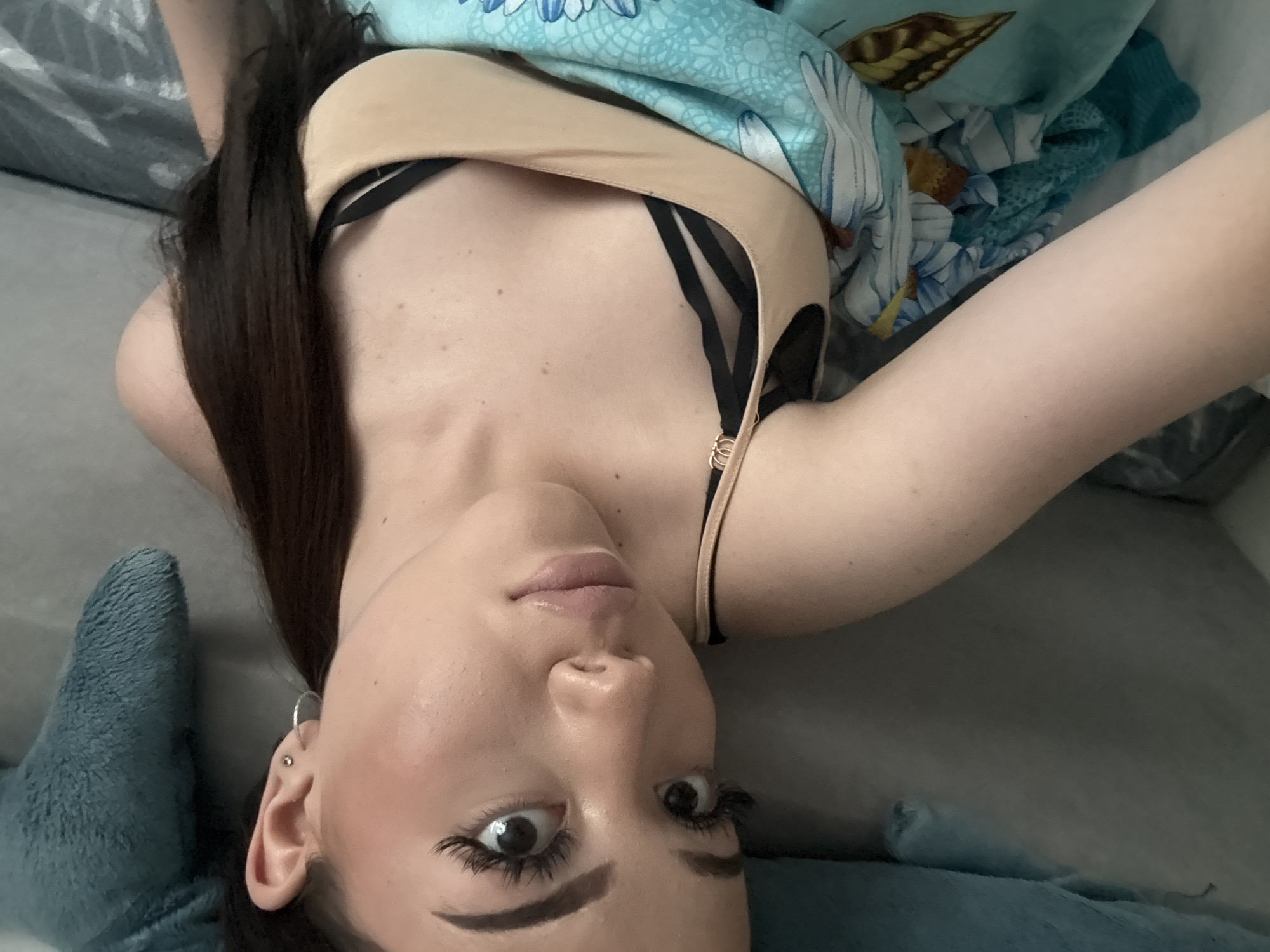 A woman is lying on a gray surface, presumably a bed, and holding a phone or camera to take a selfie. She is angled in the frame with her head at the bottom and her body extending upwards. She has dark hair, fair skin, and is wearing a tan top with black straps. A blue floral-patterned blanket or sheet is partially covering her. The environment appears to be indoors. The image does not contain enough information to determine a specific location in Naberezhnye Chelny, Russia.