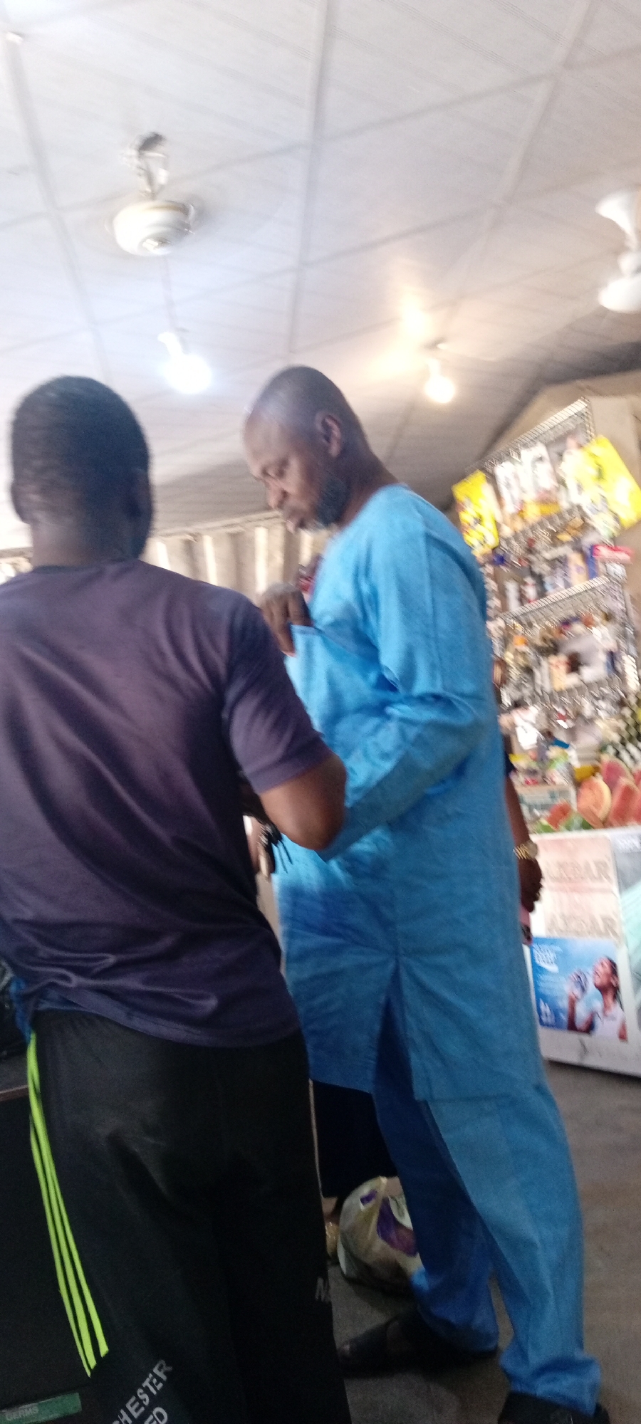 Two men are standing in what appears to be an indoor market or shop. The man in the foreground is wearing a dark purple t-shirt and black pants with neon green stripes. He is facing away from the camera. The man in the background is wearing a light blue, long-sleeved tunic and dark pants. He is looking down and appears to be reaching into his pocket. Behind him, shelves are stocked with various items, including food products and packaged goods. The ceiling has fluorescent lighting and a ceiling fan. The floor appears to be tiled or concrete. The overall lighting suggests it is daytime. There is some visible text on the pants of the man in the foreground which reads "MANCHESTER UNITED".