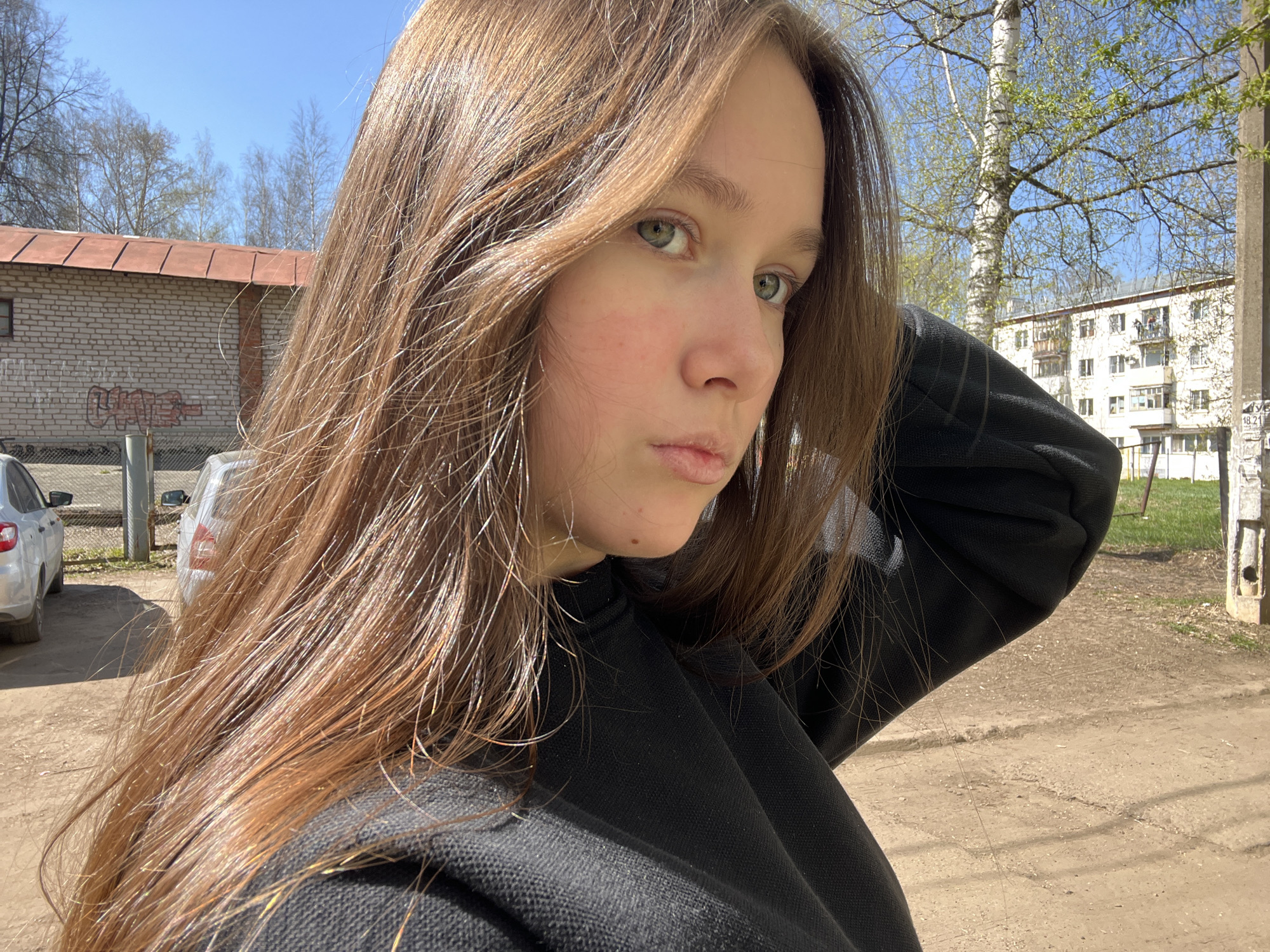 A young woman with long brown hair and light eyes stands outdoors, wearing a black hooded sweatshirt. She is positioned in the foreground, angled to the left. Behind her is a cityscape setting. A light brick building with a red metal roof is visible to the left, behind parked cars. A larger, white apartment building can be seen further back, with trees showing signs of spring. The ground appears to be a dirt or gravel surface. The image suggests it was taken on a sunny day. The location is Chaikovsky, Russia, based on the analysis.