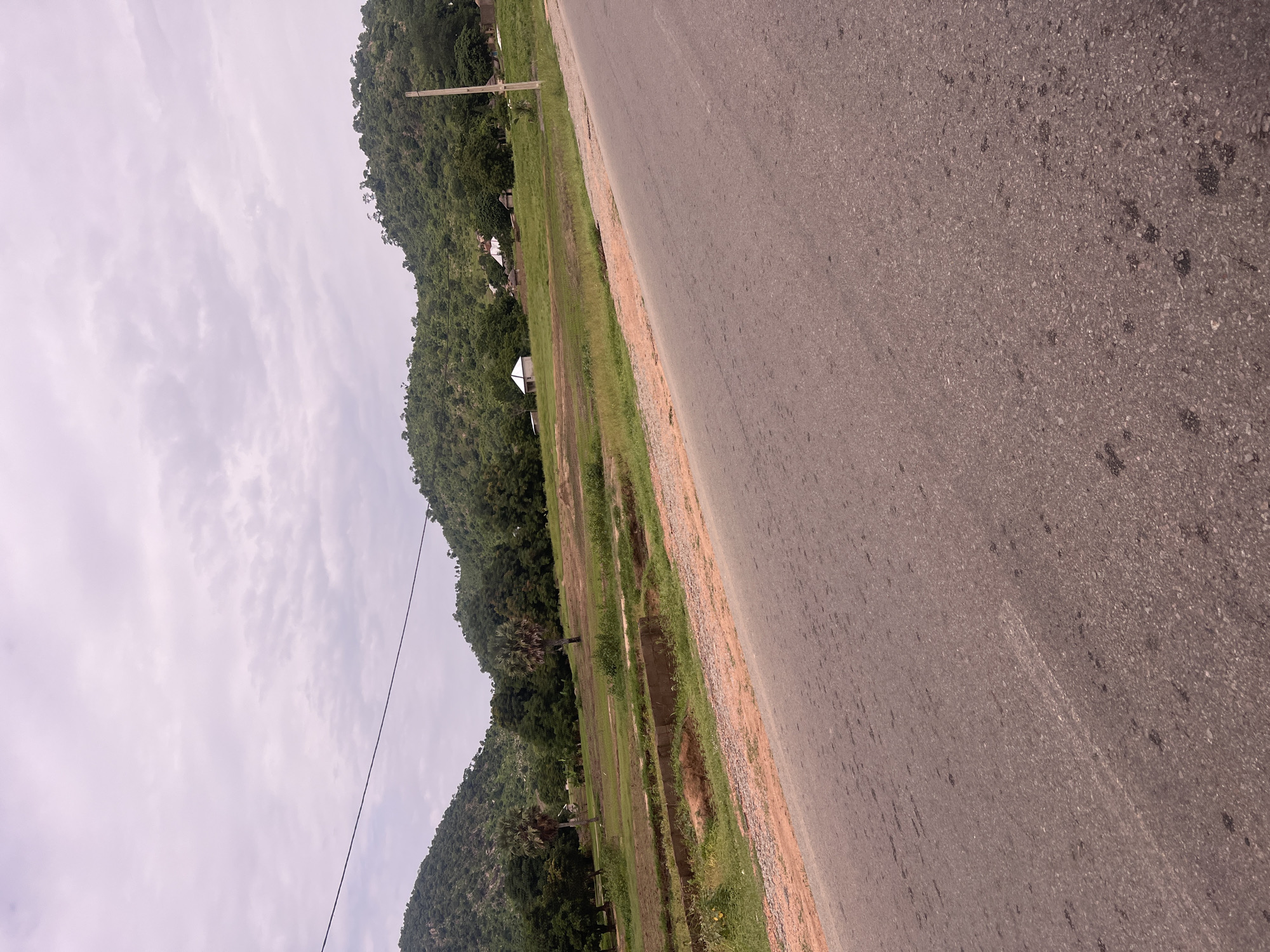An image of a rural landscape in Minda, Nigeria, features a paved road in the foreground that curves to the right, leading into the distance. Beyond the road, a grassy field stretches towards a backdrop of lush green hills and trees. Several scattered structures, including a small white building with a peaked roof and some thatched-roof huts, are visible on the lower slopes of the hills. A single utility pole stands on the right side of the frame, and a power line runs diagonally across the sky. The sky is overcast with light clouds. The scene appears calm and undeveloped, with no people or animals present.