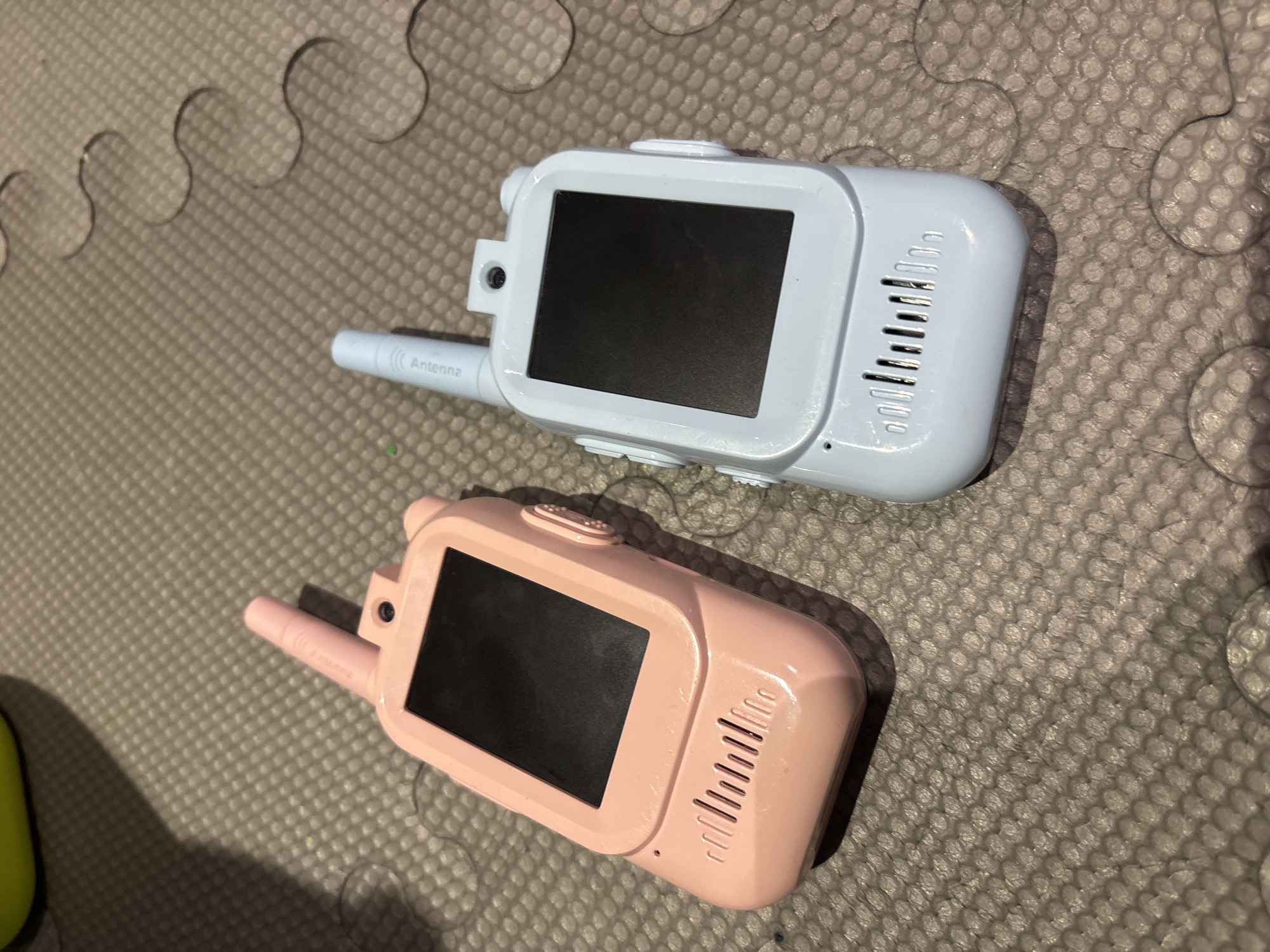 Two walkie-talkies, one pink and one light blue, lie on a textured grey mat. The pink walkie-talkie is positioned to the left, featuring a black screen, a camera, and speaker vents. The light blue walkie-talkie is to the right, also with a black screen, a camera, speaker vents, and text on its antenna that reads "Antenna". The background is indistinct, with a hint of a yellow object in the upper left corner and dark circular shapes on the mat. The image was taken indoors, likely in a residential setting. There are no people present in the image. The overall scene depicts an indoor arrangement of two children's toy walkie-talkies. The location is not discernible from the image.