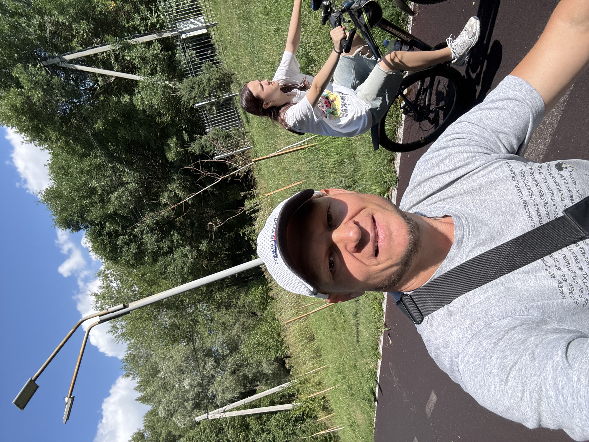 In a sunny outdoor scene, a man in a grey t-shirt and a white baseball cap is taking a selfie while riding a bicycle. Behind him, a young woman is also riding a bicycle. The road they are on appears to be a paved bike path. The background consists of lush green trees and vegetation, with a metal fence visible in the distance. Power lines and a tall lamp post are also present. The man's t-shirt has text and graphics printed on it, and the woman is wearing a white t-shirt and denim shorts. The location appears to be an urban park or recreational area in Oskemen, Kazakhstan.