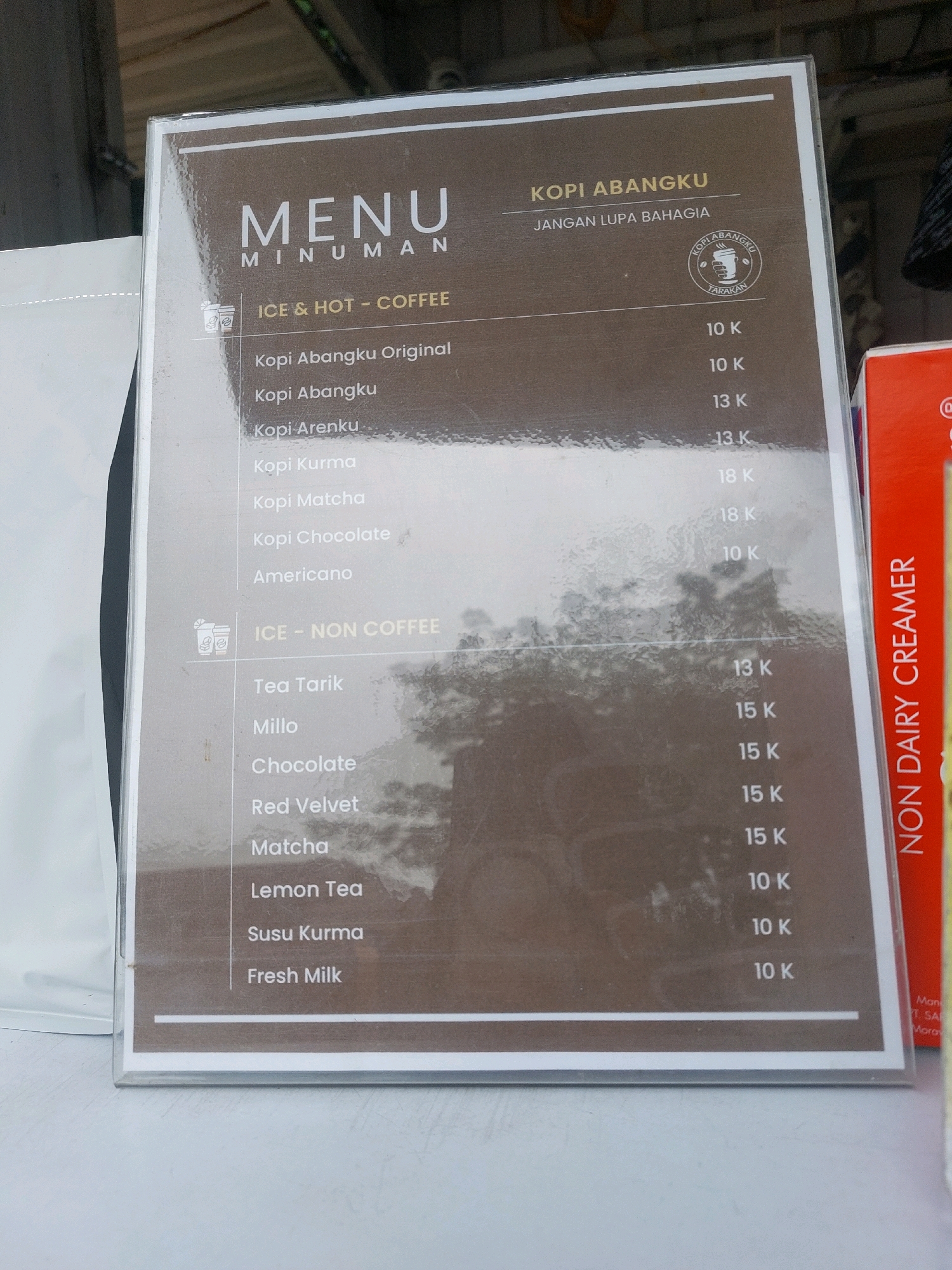 The image displays a menu board for "KOPI ABANGKU" located in Tarakan, Indonesia. The menu lists various coffee and non-coffee drinks with their prices. The items are categorized under "ICE & HOT - COFFEE" and "ICE - NON COFFEE". Specific coffee options include Kopi Abangku Original, Kopi Abangku, Kopi Arenku, Kopi Kurma, Kopi Matcha, Kopi Chocolate, and Americano. Non-coffee options include Tea Tarik, Millo, Chocolate, Red Velvet, Matcha, Lemon Tea, Susu Kurma, and Fresh Milk. The prices are indicated in thousands of Indonesian Rupiah (K). The menu board is enclosed in a plastic sleeve, and a portion of a red box with text "NON DAIRY CREAMER" is visible on the right side of the image. The background suggests an outdoor or semi-outdoor setting, possibly a food stall or cafe.
