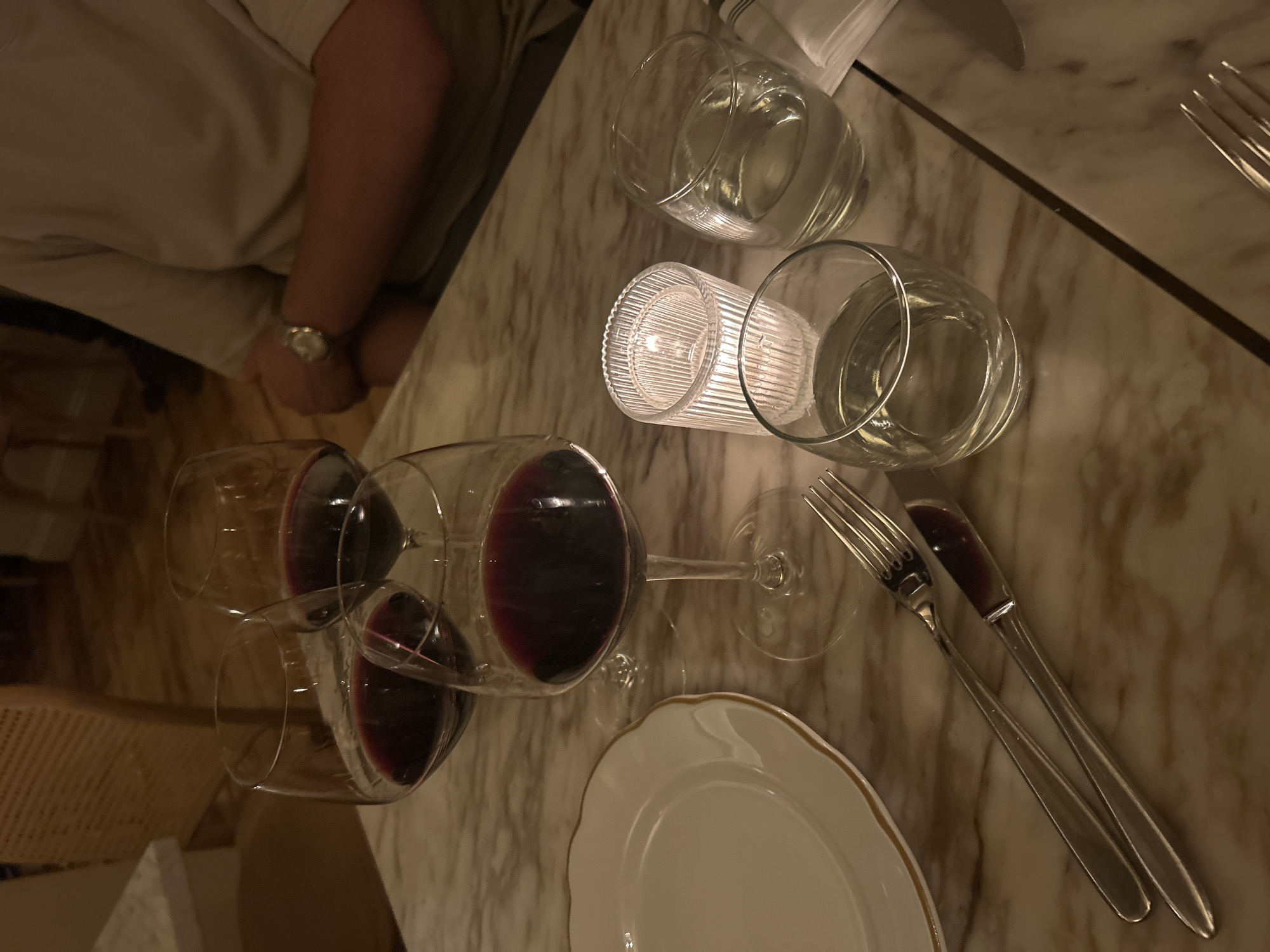 This image captures a close-up, high-angle view of a dining table at what appears to be an upscale restaurant or dining establishment in New York, United States of America, likely during the evening. The table is made of polished light-colored marble with visible veining.
In the upper left quadrant, the arm and leg of a person are visible, wearing light-colored clothing (possibly a white shirt and cream trousers) and a wristwatch with a metallic strap on the left wrist, indicating the presence of at least one diner.
The table setting includes several clear glass items: two tumblers filled with water, and two stemmed wine glasses, both containing red wine. A ribbed glass candle holder, emitting a soft glow, is centrally placed on the table, contributing to the intimate and warm ambiance. Adjacent to it, another empty clear glass is visible.
Towards the bottom right, a white round plate with a delicate gold rim is set, accompanied by a silver fork and knife arranged diagonally to its upper right. A folded white napkin is also partially visible in the upper right corner.
The lighting is dim, suggesting a nighttime setting, and the glow from the candle is a prominent light source, casting soft reflections on the glasses and the table surface. No text is visibly present in the image. The overall scene suggests a sophisticated and relaxed dining experience.