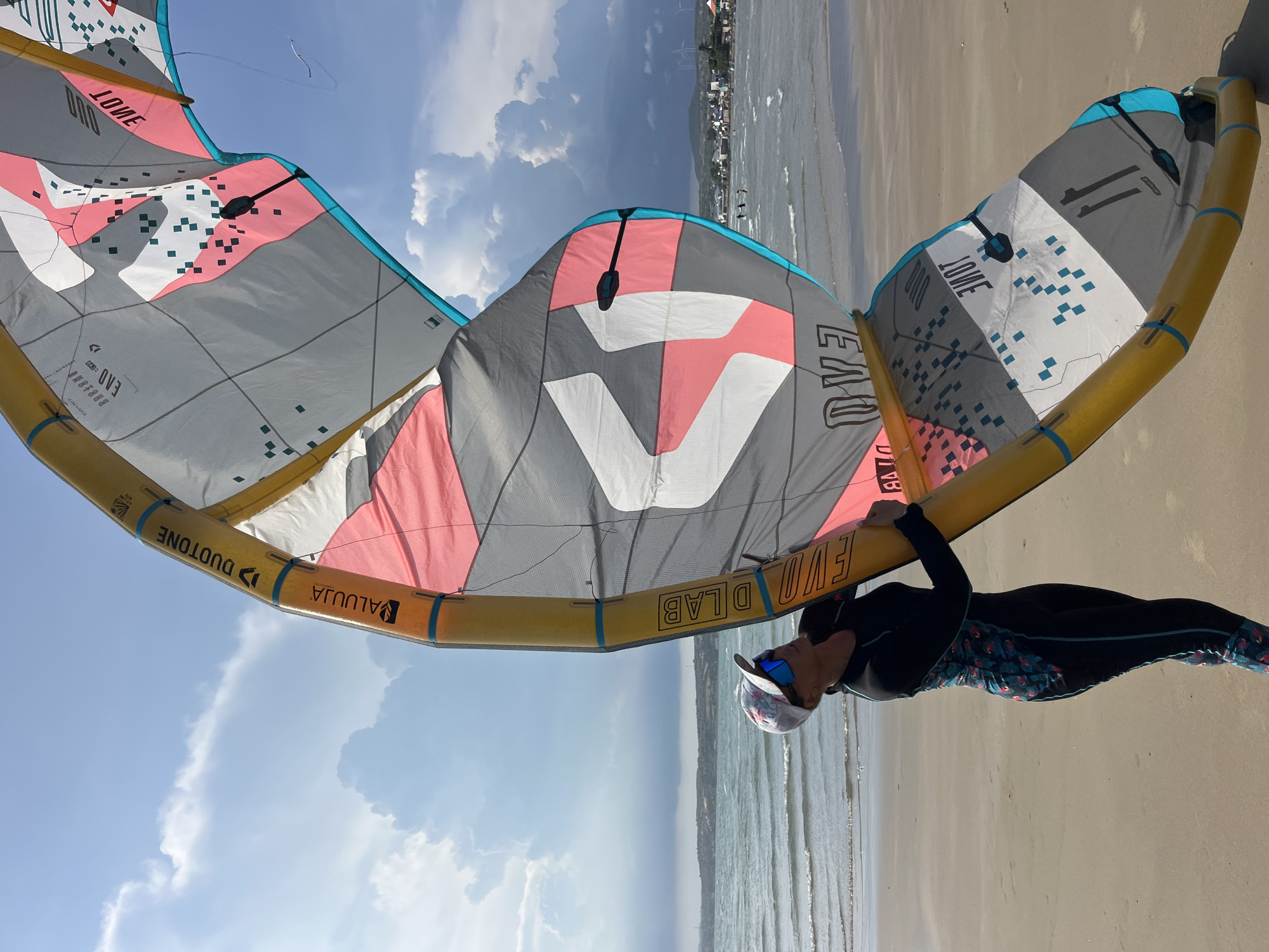 A woman, wearing a black and patterned wetsuit, a white baseball cap, and blue mirrored sunglasses, stands on a sandy beach in Mũi Né, Vietnam, holding a large, yellow-framed kite. The kite is predominantly gray, white, and pink, with teal accents and square patterns. The kite is partially inflated and angled upwards, obscuring some of the sky and background. The woman is looking up and slightly to her left, her hands on the kite's frame.
The background features a calm ocean with gentle waves, a distant shoreline with a town and wind turbines, and a cloudy sky with patches of blue. Several people are visible in the water in the distance, appearing small against the expanse of the sea. The overall lighting suggests a bright, daytime scene.