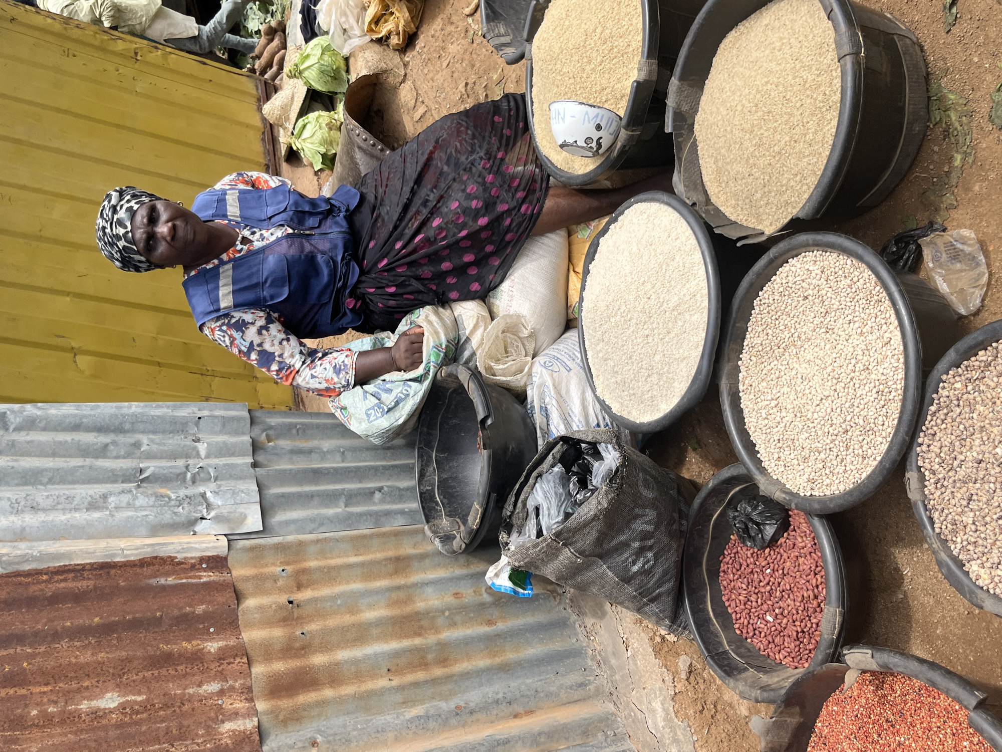 In Gura Top, Nigeria, a woman in a blue vest and a headscarf sits behind a market stall overflowing with various grains. She is positioned amidst several large black buckets, each filled to the brim with different types of grains: red beans, white rice, and a lighter, tan-colored grain are clearly visible. Behind her, a large bag of grain rests on a white sack. The stall's backdrop consists of corrugated metal sheeting, some of which is rusted, and a bright yellow structure. In the background, glimpses of other market-goers and produce like cabbages and yams suggest a busy marketplace environment. The ground is a mix of dirt and sand.