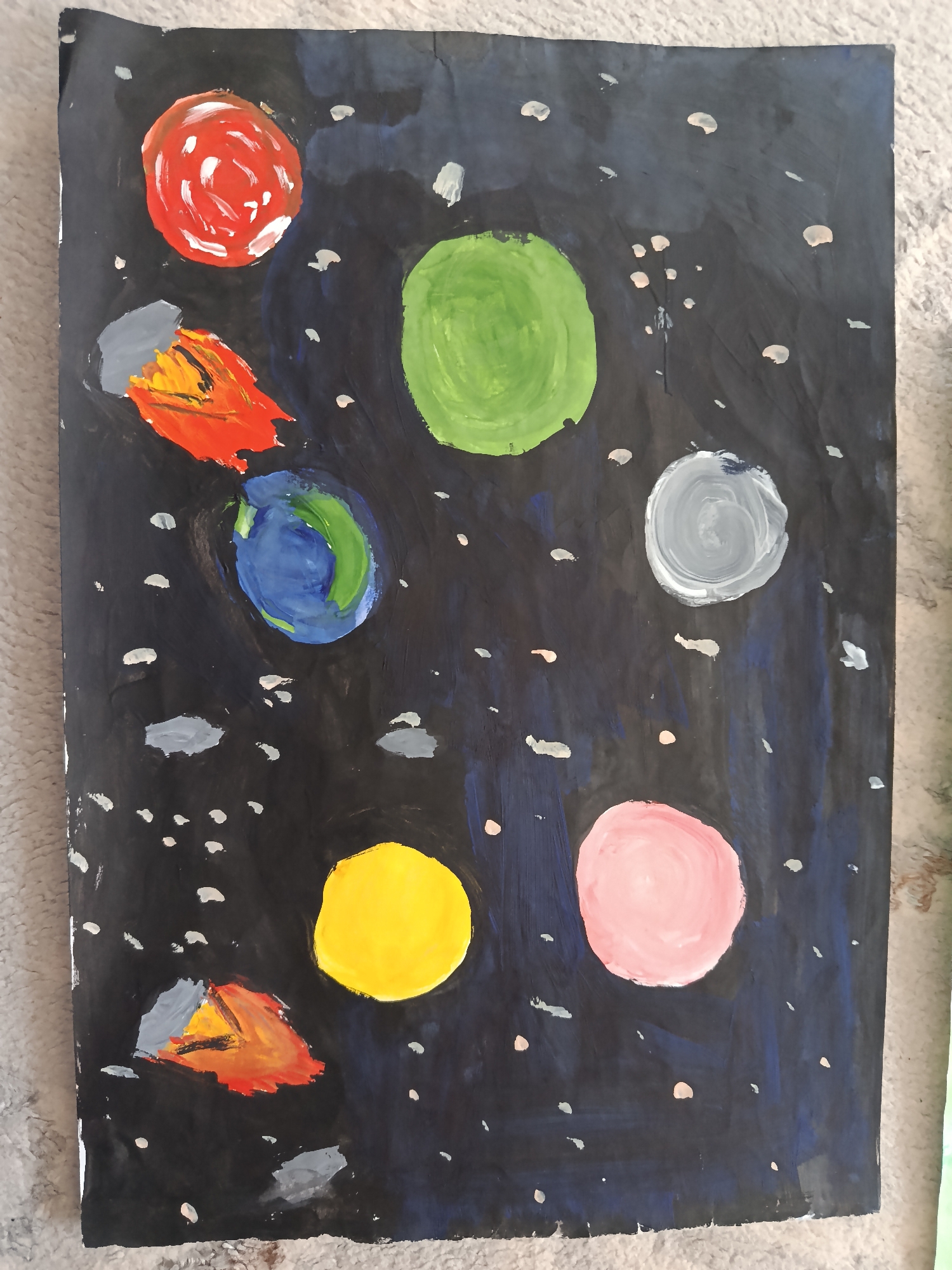 An acrylic painting on black paper depicts a starry night sky with various celestial bodies. A red, circular object is in the upper left, with a streak of orange and yellow originating from its lower left. Below this, a blue and green orb represents Earth. To the right of Earth is a large green circle, followed by a gray and white swirling circle further to the right. In the bottom left, another orange and yellow streak emanates from a gray triangular shape. Below that, a bright yellow circle dominates the lower middle of the painting. To the right of the yellow circle is a large pink circle. Numerous small white dots are scattered across the black background, suggesting stars. The painting is slightly crumpled at the edges.