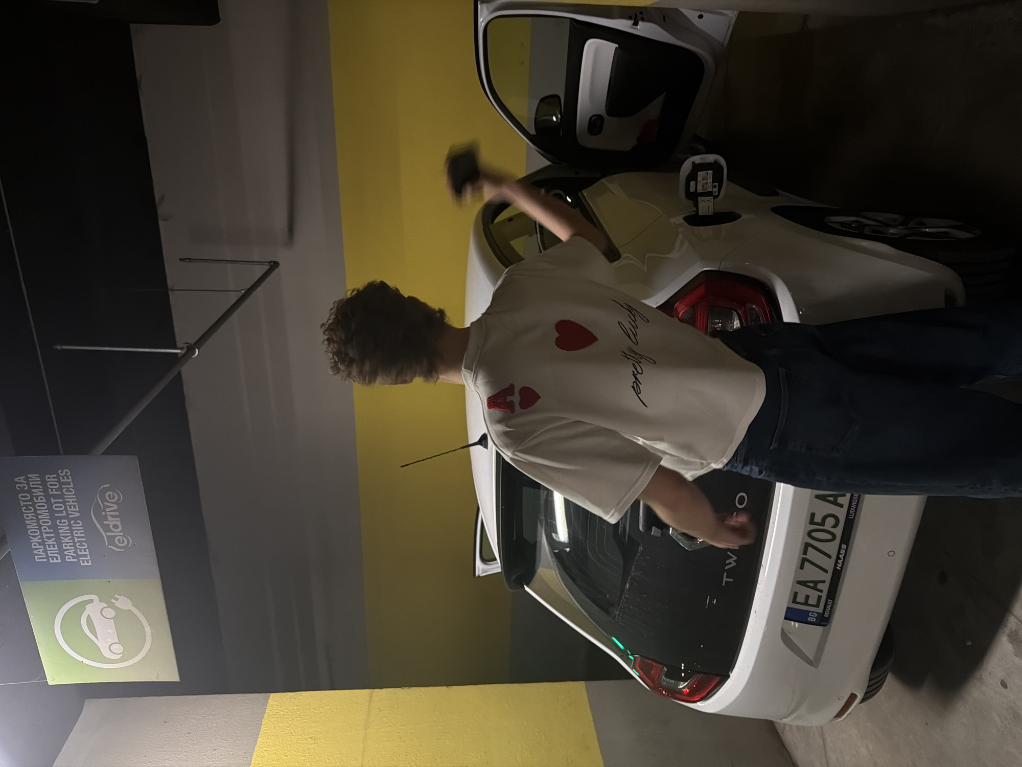 An indoor, portrait-oriented photograph depicts a person interacting with a white electric car in a dimly lit parking garage. The subject, seen from behind and to the side, has curly brown hair and is wearing a white t-shirt adorned with red heart graphics and the text "pretty Judy." They are dressed in dark jeans and appear to be handling a charging cable, with the car's charging port open.
The car is a white Renault Twizy, partially visible, with its rear license plate "BG EA 7705 A" clearly displayed, indicating its origin from Bulgaria. The license plate frame also shows "RENAULT", "HAASS", and "LUDWIGSE". The vehicle is parked next to a wall section painted in dark grey, light grey, and yellow stripes.
To the left of the car, a sign is visible, written in both Cyrillic and English, stating "ПАРКОМЯСТО ЗА ЕЛЕКТРОМОБИЛИ / PARKING LOT FOR ELECTRIC VEHICLES" and featuring the "eldrive" logo, confirming the car's electric nature and the designated parking spot. The scene suggests the charging of an electric vehicle in a public or private underground parking facility, likely at night or in low light conditions, in Sofia, Bulgaria.
