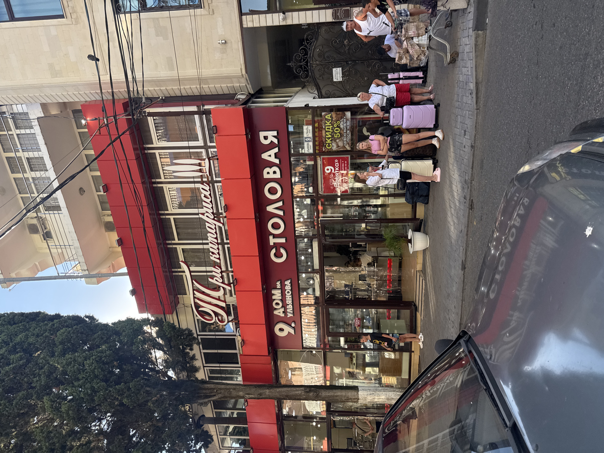 A street scene in Sochi, Russia, features a red-fronted building with large glass windows displaying merchandise, possibly a store or a cafe. A sign above the entrance reads "9 ДОМ НА УЛЬЯНОВА СТОЛОВАЯ" in large white letters. To the left, a tall green tree stands beside the building. Several people are gathered on the sidewalk in front of the establishment, some with luggage, suggesting they are travelers. One person is leaning against the entrance. A dark gray car is parked in the foreground, partially obscuring the view of the sidewalk. The street is paved with gray cobblestones, and a few potted plants are visible. The overall impression is a commercial street in a city, possibly near a transport hub or tourist area.