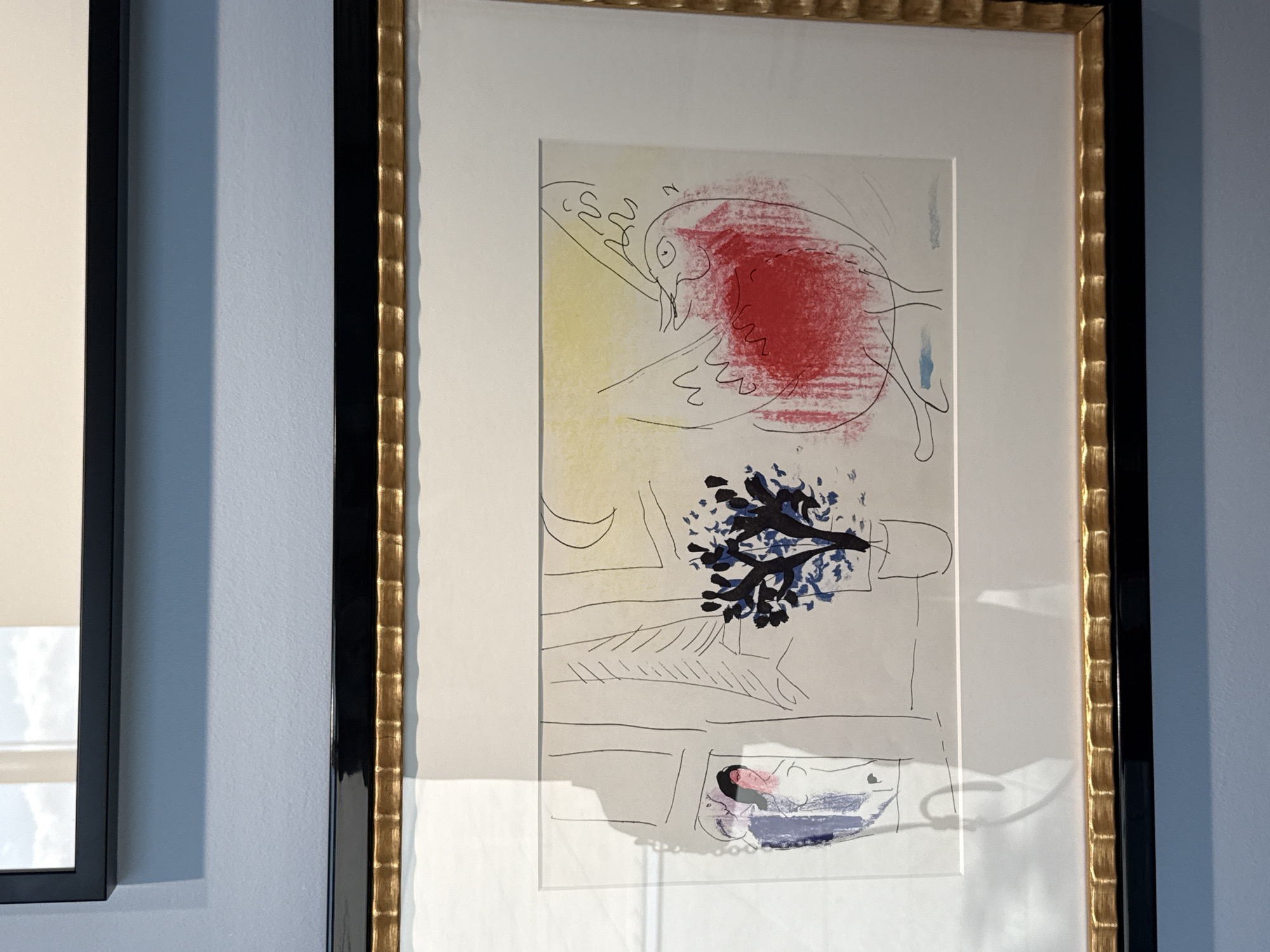 The image displays a framed artwork hanging on a light blue wall. The artwork itself is a drawing with watercolor elements, framed by a black mat and a textured gold and black frame. The scene depicted in the artwork includes a stylized bird with a large red body, a woman in a doorway or window, a potted plant with dark blue foliage, and a crescent moon in a yellow sky. The style is abstract and somewhat whimsical. A shadow is cast across the left side of the artwork, obscuring a portion of the woman and the background. The room appears to be an interior space, possibly a gallery or home, given the artwork and wall color.