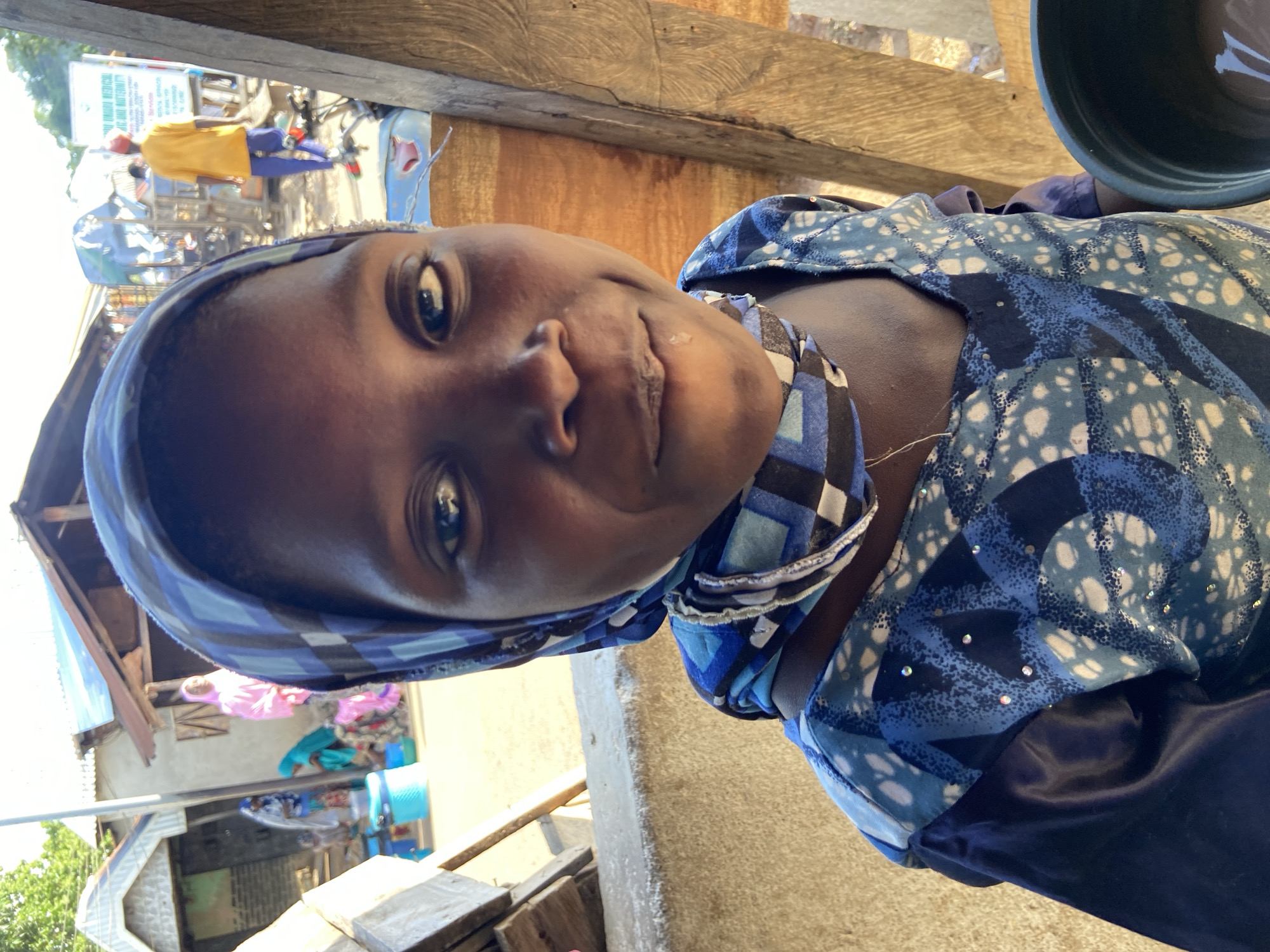 A medium shot of a person with dark skin and dark eyes wearing a blue and white patterned headscarf and matching top in an outdoor setting. The subject is smiling slightly at the camera. A black pot is visible in the top right corner of the frame. Behind the subject, there appears to be a street scene with buildings, possibly located in Mararaba, Nigeria, as evidenced by the presence of local structures and activity in the background.