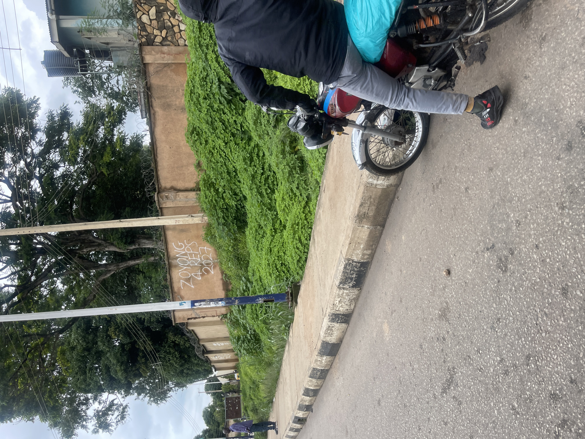 The image shows a street scene in Jos, Nigeria. In the foreground, a person is seated on a red motorcycle, their back to the camera. They are wearing a black jacket and light gray pants. The motorcycle has a bright blue tarp covering the rear seat. The motorcycle's front wheel and handlebars are visible.
The street itself is asphalt, with some dark spots and a single small rock in the center. To the left of the road is a concrete sidewalk with black and white striped edging. A tall, white utility pole with electrical wires runs along the sidewalk.
Behind the sidewalk, a concrete wall lined with barbed wire runs along the street. The wall is plastered and painted a tan color, with white spray paint graffiti that reads "ZOYONG 4 REPS 2027". Above the wall, dense green vegetation covers a slope. Further back, trees and a building with a water tank are visible against a cloudy sky.
To the far left of the image, a man in a plaid shirt and dark pants stands on the sidewalk, facing away from the motorcycle. He appears to be looking at something off-camera. The overall impression is a typical urban street environment in Jos.