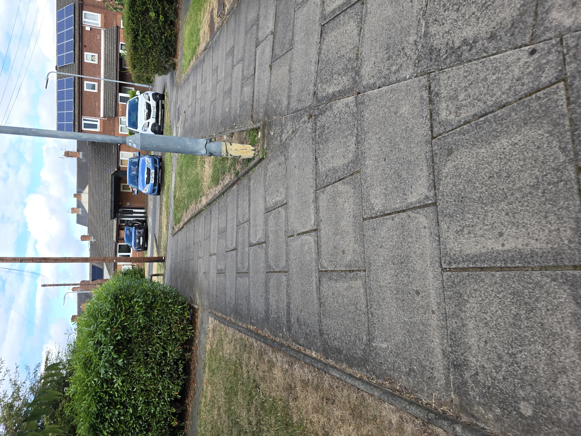 The image shows a paved footpath in Stockton-on-Tees, United Kingdom, with a large hedge on the left and a row of houses with solar panels on the roof on the right. Two cars, a blue Skoda Fabia and a white Vauxhall Corsa, are parked in front of the houses. A street lamp is visible in the middle of the frame. The ground is paved with large concrete slabs, and there is dry grass on either side of the path. The sky is blue with some clouds.