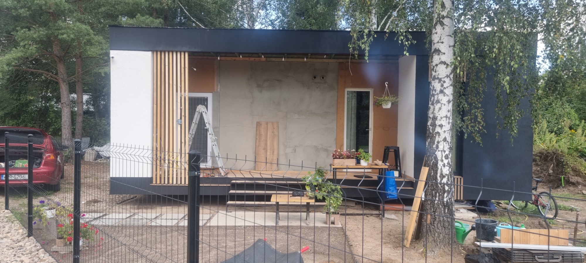 The image depicts an exterior view of a partially constructed modern house in Szczaki, Poland. The house has a black roofline and a mix of white, natural wood, and grey concrete exterior walls. A large sliding glass door is visible on the right side of the house, and a standard door with a metal ladder leaning against it is on the left. There are wooden slats on the left side of the structure, and the right side is covered in dark material. In front of the house, a wooden deck with stairs leads to the entrance. A black metal fence encloses the property, and a red car is parked on the left side. Various construction materials, gardening tools, and potted plants are scattered around the scene, suggesting ongoing work. A birch tree stands prominently to the right of the house. The environment is natural and appears to be a rural or semi-rural area with trees and vegetation surrounding the property.