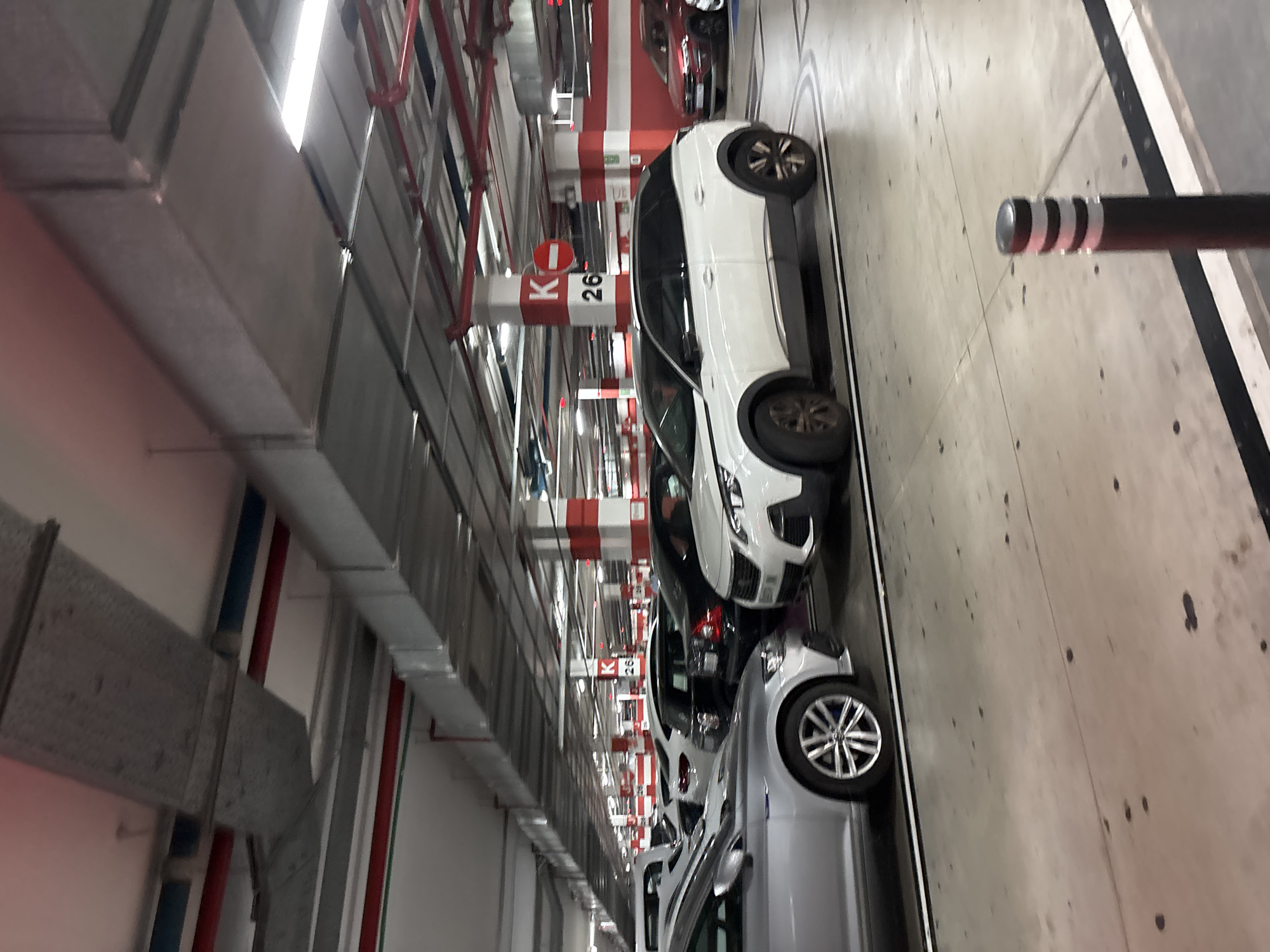 The image depicts a section of an underground parking garage in Orihuela, Spain. In the foreground, a silver car is parked on the left, and a white SUV is parked on the right. Behind them, other cars are visible, including a black hatchback and a red sedan further in the background. The parking garage features concrete pillars painted with alternating red and white bands, and signage indicating "K 26". Overhead, metal ductwork and lighting fixtures are mounted on the ceiling. The floor is concrete with painted lines defining parking spaces. A black bollard with reflective white bands stands near the foreground. The overall scene is static, showing a typical environment within a parking facility.