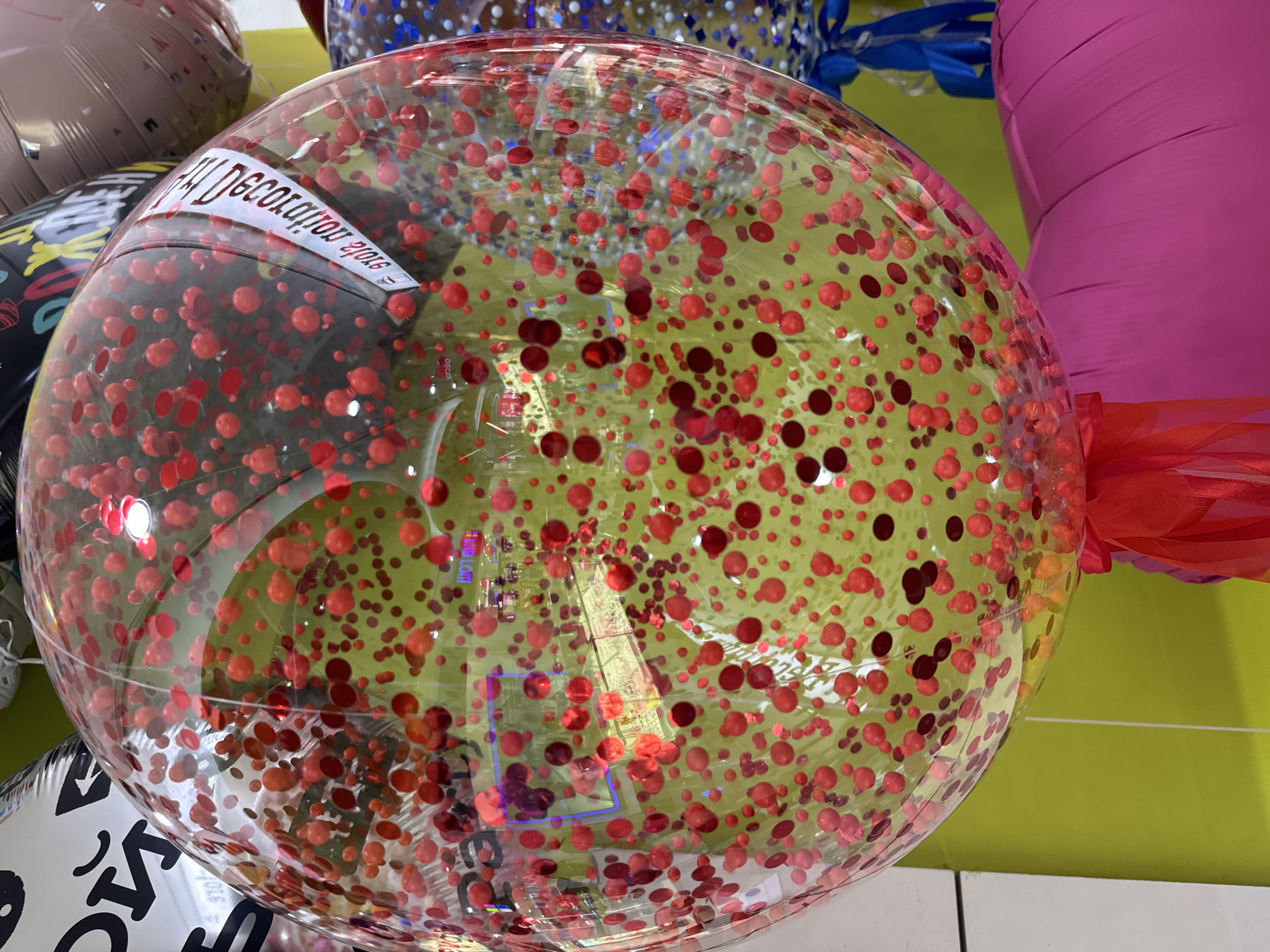 A close-up, slightly tilted shot captures a large, clear balloon filled with red confetti. The balloon is adorned with a white sticker that reads "GM Decoration". Behind the main balloon, parts of other balloons are visible, including one with a black and white patterned design and another in a metallic brown hue. Below the balloon, a bright pink inflatable object is partially visible. The background features a lime green wall and a glimpse of what appears to be a retail store interior with display shelves. The scene is well-lit, possibly in a shop in Sinyavino, Russia.
