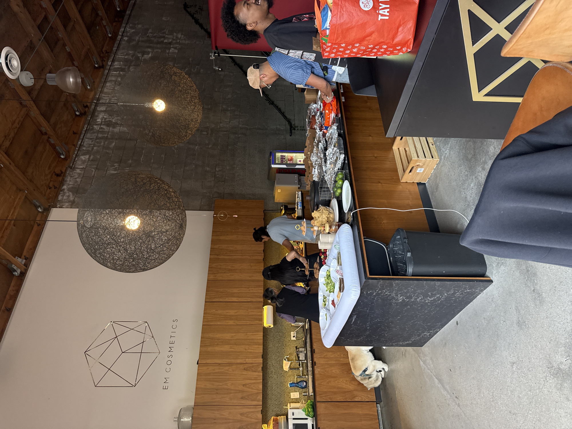 This image shows the interior of a business, likely a restaurant, in Culver City, California. Two large, spherical pendant lights hang from the ceiling. A counter with multiple people preparing food stretches across the length of the image. The counter is lined with trays containing various ingredients. A dog is lying on the floor near the end of the counter. Customers are standing by the end of the counter, with one holding a red shopping bag. A sign with "EM COSMETICS" is visible on a white wall next to a wooden cabinet.