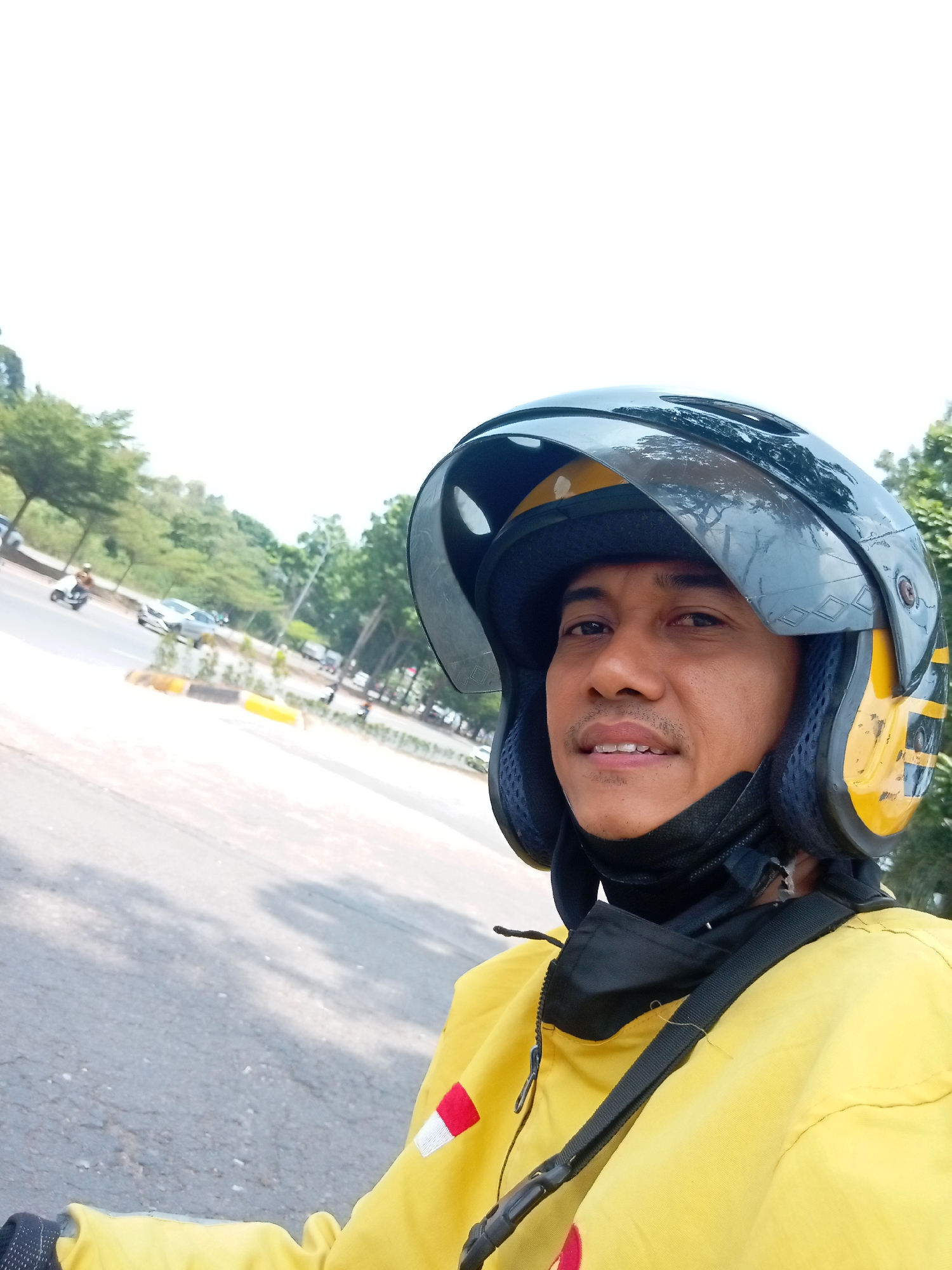 A man wearing a yellow jacket and a black and yellow helmet is captured in a close-up selfie. He is on a road with trees and a few cars in the background, suggesting an outdoor urban setting. A red and white flag patch is visible on his left sleeve. The man appears to be in motion, possibly riding a motorcycle, given his attire. The scene is likely in Batam, Indonesia, a city known for its urban landscapes and transportation.