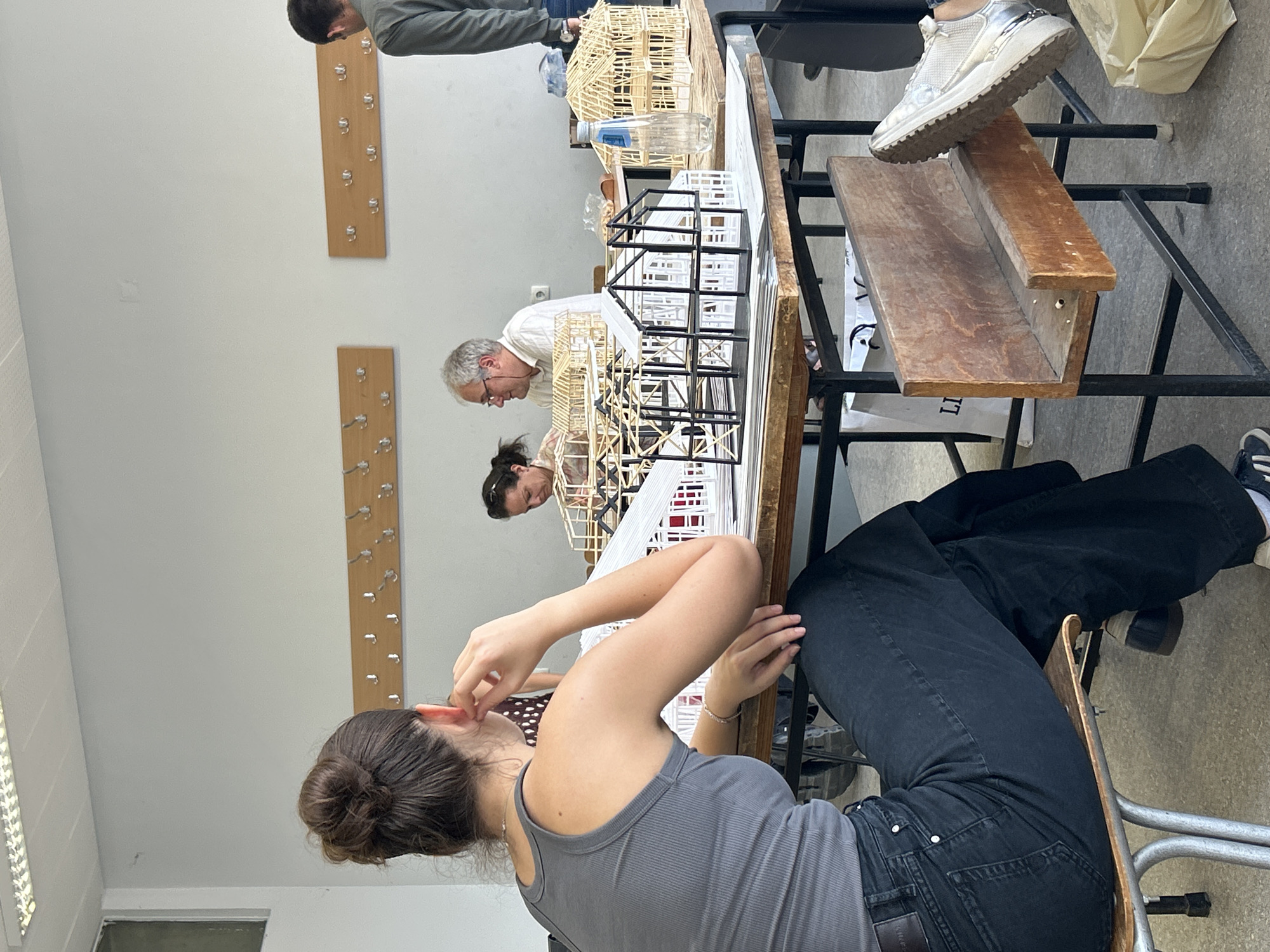 The image depicts an indoor scene, likely a classroom or workshop, where individuals are engaged with architectural models. In the foreground, a young woman with her hair pulled back in a bun, wearing a grey tank top and dark, wide-legged pants, sits with her back mostly to the camera, resting her hand on her ear. She is seated at a wooden table where several architectural models are displayed.
On the table, various models are visible. Some are intricate structures made of thin wooden sticks, resembling houses or buildings with exposed framing and roof structures. There are also models with black frames and white infill, presenting a more modern architectural aesthetic. A clear plastic bottle of water is placed on the table near one of the wooden models.
Further back in the scene, two individuals are seated at another table. A woman with dark hair and glasses is looking towards the models, and a man with grey hair and glasses is also looking in the same direction, appearing engaged with the architectural designs. Another person, partially visible on the right edge of the frame, is standing and wearing a grey long-sleeved shirt.
The environment includes walls with light-colored paint, adorned with two wooden racks holding multiple coat hooks. Fluorescent lighting fixtures are visible on the ceiling. The floor appears to be concrete. The overall impression is one of an academic or creative setting focused on architectural model building. The specific location in Sofia, Bulgaria, is not identifiable from the visual elements within the image.