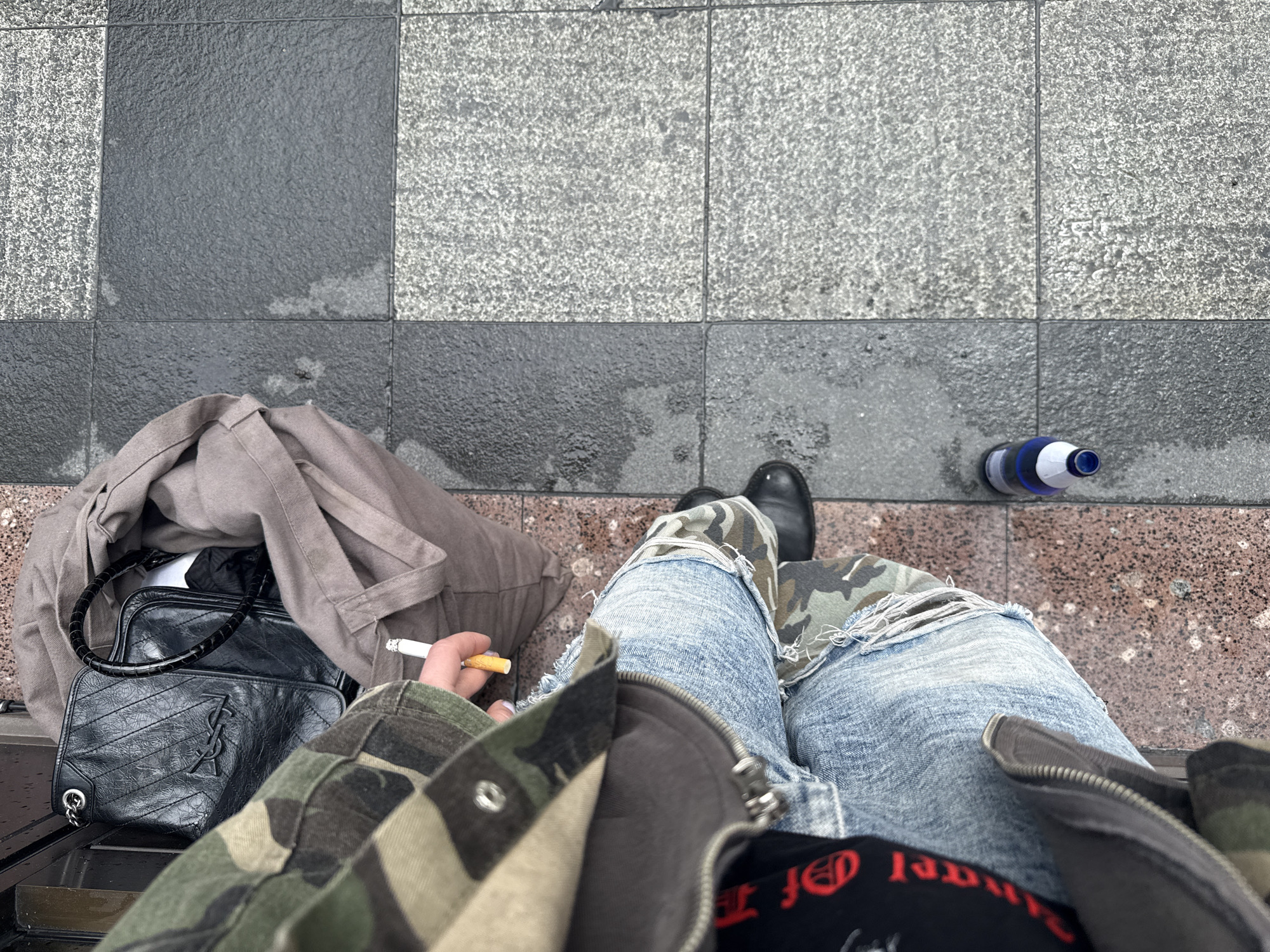 A first-person, overhead perspective captures a scene on a possibly wet, tiled surface with varied textures, appearing to be outdoors. The lower half of a person is visible, wearing ripped, light-wash jeans and black leather shoes. They are seated, knees slightly bent, and holding a lit cigarette in their left hand. A camouflage jacket, partially zipped, is visible on the person's upper body, revealing a black T-shirt with red lettering underneath. To the left of the person, a black designer handbag with a "YSL" logo sits beside a folded beige jacket or bag. A partially visible blue water bottle lies to the right of the person's right shoe. There is no indication that the location is in Sochi, Russia.
