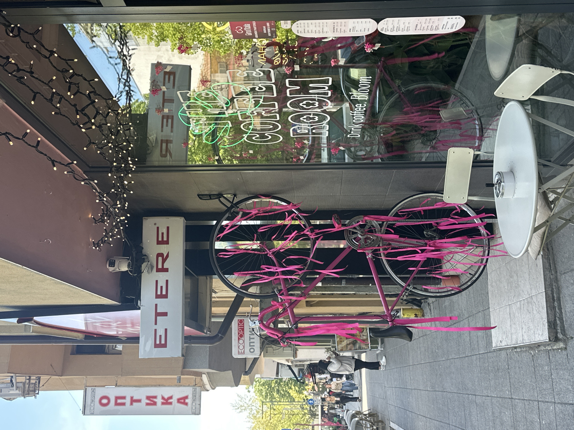 Here is a detailed description of the image:
The image shows a street scene, likely in Burgas, Bulgaria, based on the Cyrillic script present on the signs. The scene is captured from a street-level perspective, looking at a storefront. A pink bicycle decorated with pink ribbons is mounted on the wall above the storefront. The store's window displays reflective signs saying "COFFEE ROOM" and "Drink coffee. Bloom", as well as other text in both English and Bulgarian. Above the storefront, there are commercial signs, including one that reads "ETERE" and "ОПТИКА ECOOPTIC". A string of lights is draped along the building's edge. A security camera is also visible above the storefront. Pedestrians are walking on the sidewalk, partially visible in the background. A white chair and table set are on the sidewalk adjacent to the store. The sidewalks are paved with grey tiles. The sky is visible between the buildings.