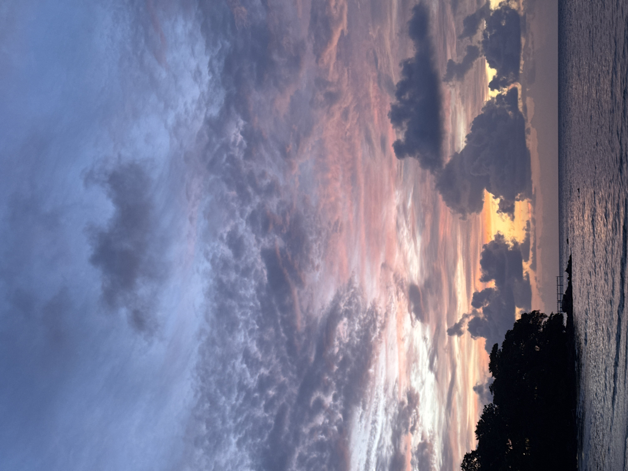 A silhouetted landmass with trees is visible on the left side of the image, jutting into a body of water. A pier or walkway extends from the land into the water. The water surface reflects the colors of the sky. The sky is filled with clouds in various shades of blue, pink, and orange, indicative of a sunset or sunrise. There are no people or animals visible in the image. The scene appears to be a coastal area, possibly within or near Patong, Thailand, given the visual characteristics of tropical foliage and the ocean.