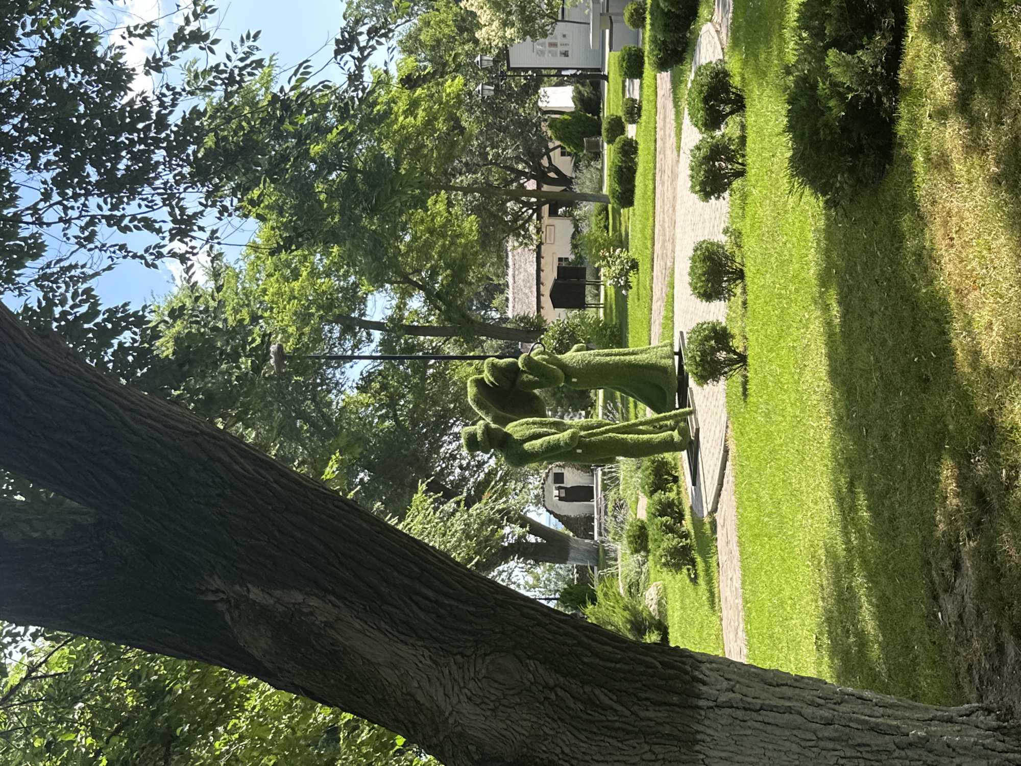 In the city of Taman, Russia, an outdoor scene is depicted in a park setting. A prominent tree trunk occupies the left foreground, casting shadows on the meticulously manicured lawn. In the middle ground, two topiary figures, crafted to resemble a man and a woman, stand near a paved pathway. The male figure wears a hat and holds a cane, while the female figure appears to have an umbrella. Beyond the topiaries, a quaint building with a thatched roof is visible, suggesting a historical or cultural point of interest. The environment is lush and green, with well-maintained bushes and trees adorning the park. The sky above is clear and blue, indicating a pleasant day.