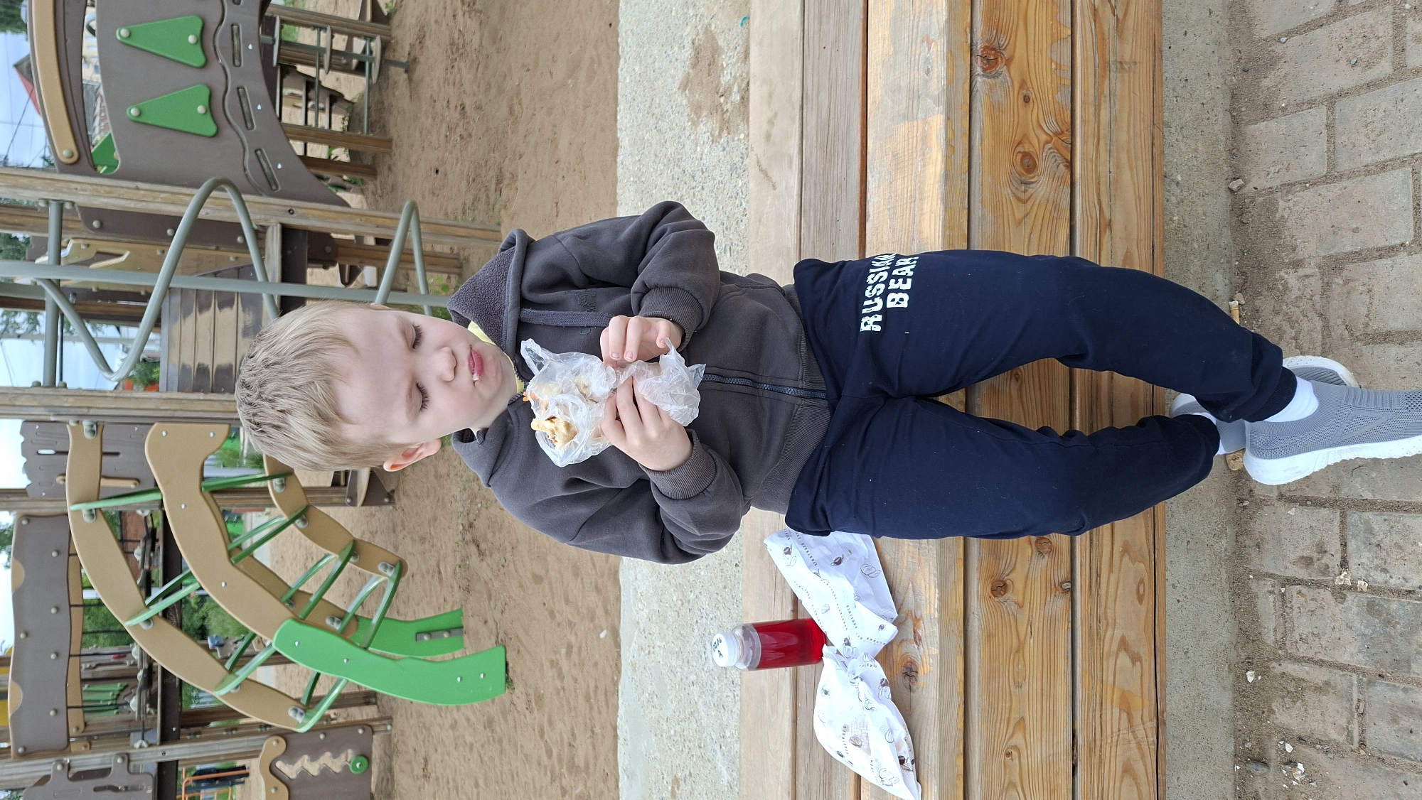 A young boy with blonde hair is sitting on a wooden bench in a playground in Nytva, Russia, eating a food item wrapped in plastic. He is wearing a dark gray hooded sweatshirt and dark blue sweatpants with "RUSSIA BEAR" printed on the left leg. He has white socks and gray sneakers. To his left on the bench are two white wrappers, one of which appears to be for a food item, and a small red bottle. The playground equipment, consisting of a colorful, multi-level structure, is visible in the background. The ground is covered in sand and brick pavers.