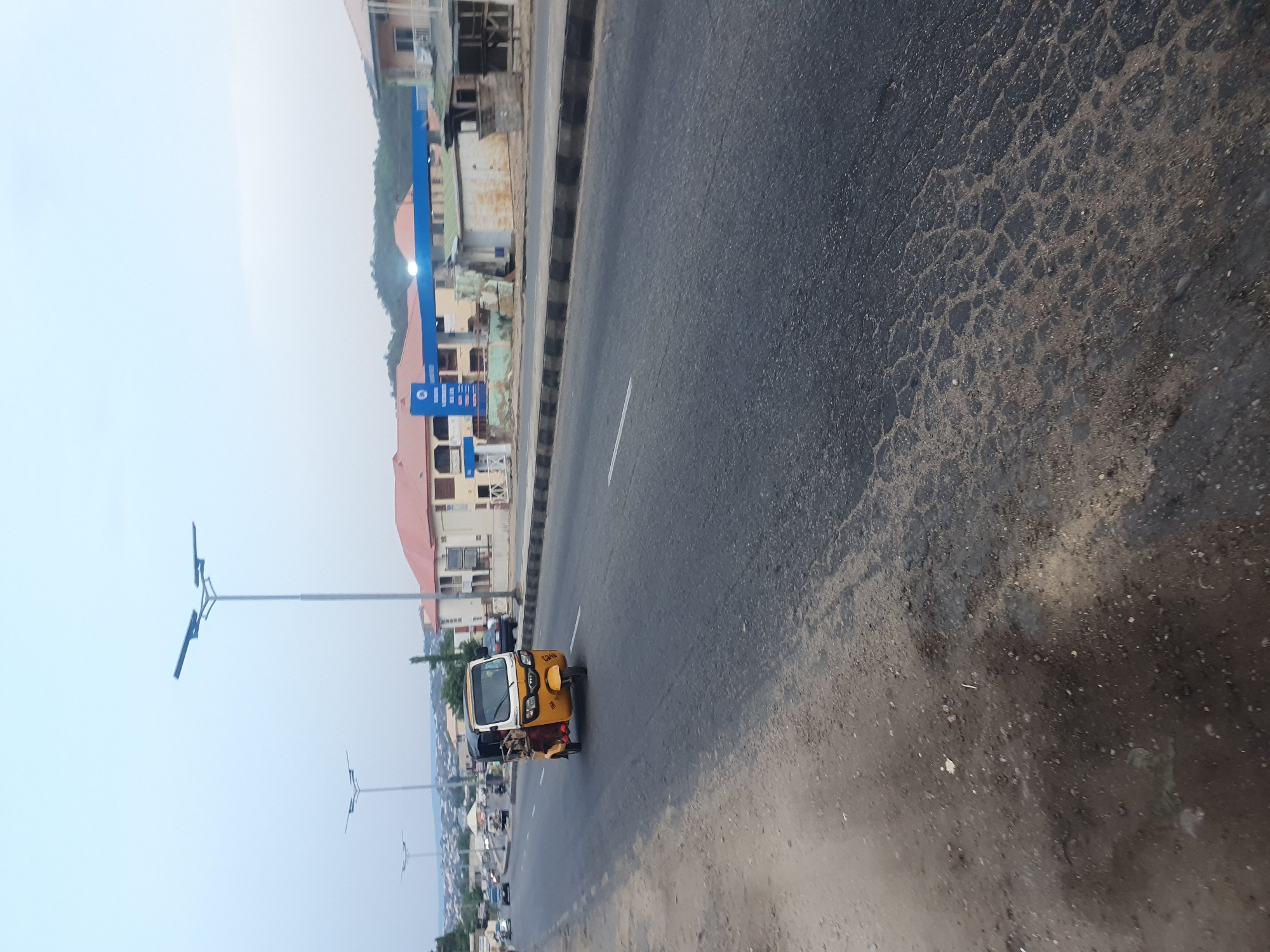 The image depicts a road in Lokoja, Nigeria, with a yellow and white tuk-tuk (auto rickshaw) traveling along the asphalt surface. The road exhibits signs of wear and tear. On the roadside, buildings with red roofs and blue signage are visible. Streetlights line the edge of the road. The sky is overcast. Other vehicles are present on the road.
