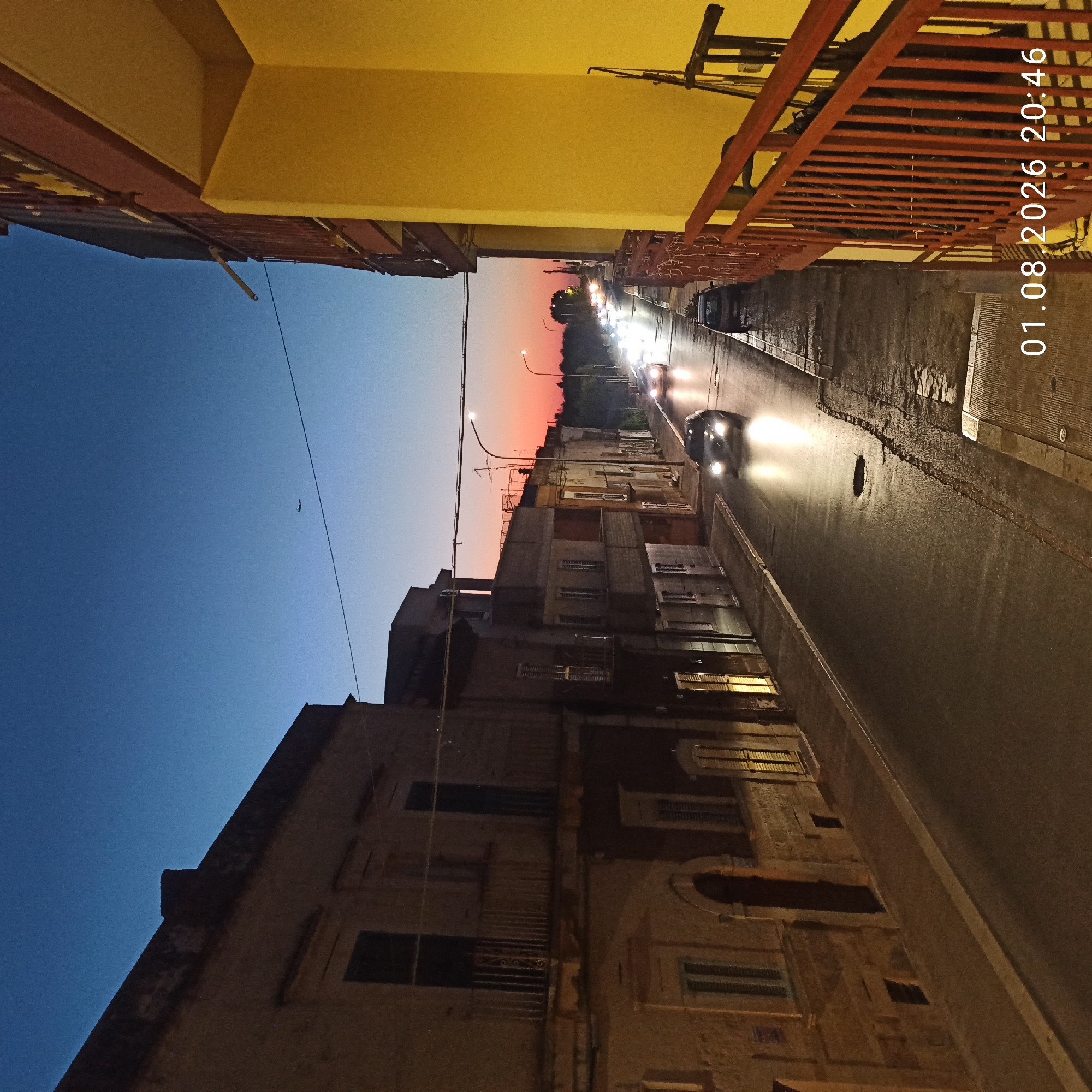 An exterior, eye-level, medium shot captures a street scene in a European city at dusk. The sky is a gradient of deep blue at the top, transitioning to pink and orange near the horizon. Buildings line both sides of the street, with the building on the left being a muted yellow and the building on the right a vibrant yellow. The street is paved with asphalt and appears wet, reflecting the headlights of passing cars. A few cars are visible on the street, moving in the distance, their headlights on. There is a small airplane in the upper portion of the sky. The image is dated and time-stamped at the bottom right corner: "01.08.2026 20:46".