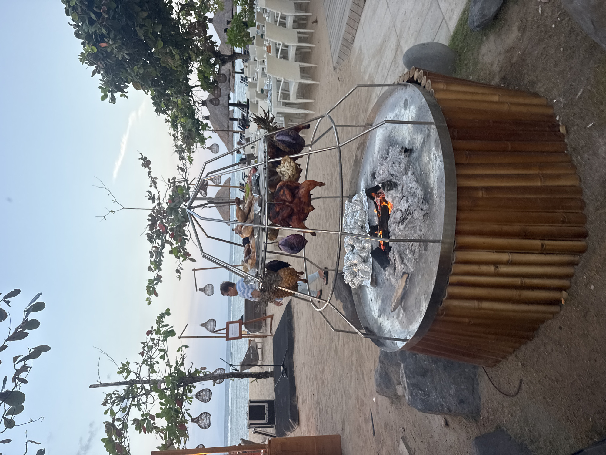 This image depicts an outdoor dining area on a sandy beach, likely in Kuta Selatan, Indonesia, as suggested by the presence of tropical vegetation, thatched-roof umbrellas, and the ocean visible in the background. The focal point is a large, circular grill with a bamboo base. Meats, including what appears to be whole chickens and possibly fish, are suspended and roasting over hot coals in the center of the grill. Beside the grill, several pineapples are also being prepared for cooking. In the background, tables and chairs are set up on the sand, suggesting a restaurant or resort setting. A person is walking by the grill, and other patrons can be seen relaxing under the umbrellas further down the beach. The sky is a soft blue, indicating either early morning or late evening.