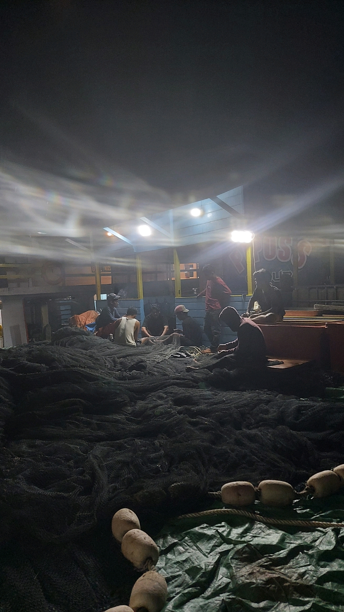 The image depicts a group of individuals, likely fishermen, gathered on what appears to be a fishing vessel at night. The scene is illuminated by artificial lights, casting long shadows and creating a somewhat hazy atmosphere. The foreground is dominated by a large pile of dark fishing nets, with some netting also spread out on a green tarp. Several buoys, attached to a rope, are visible in the lower right portion of the frame. In the background, under a covered structure with ceiling lights, several men are seated or standing, some appearing to be mending or organizing the nets. One individual in a red shirt stands prominently in the mid-ground. The overall environment suggests a working harbor or dock area in Tanjung Redeb, Indonesia, given the context. The image captures a moment of communal activity, likely preparation or post-fishing tasks.